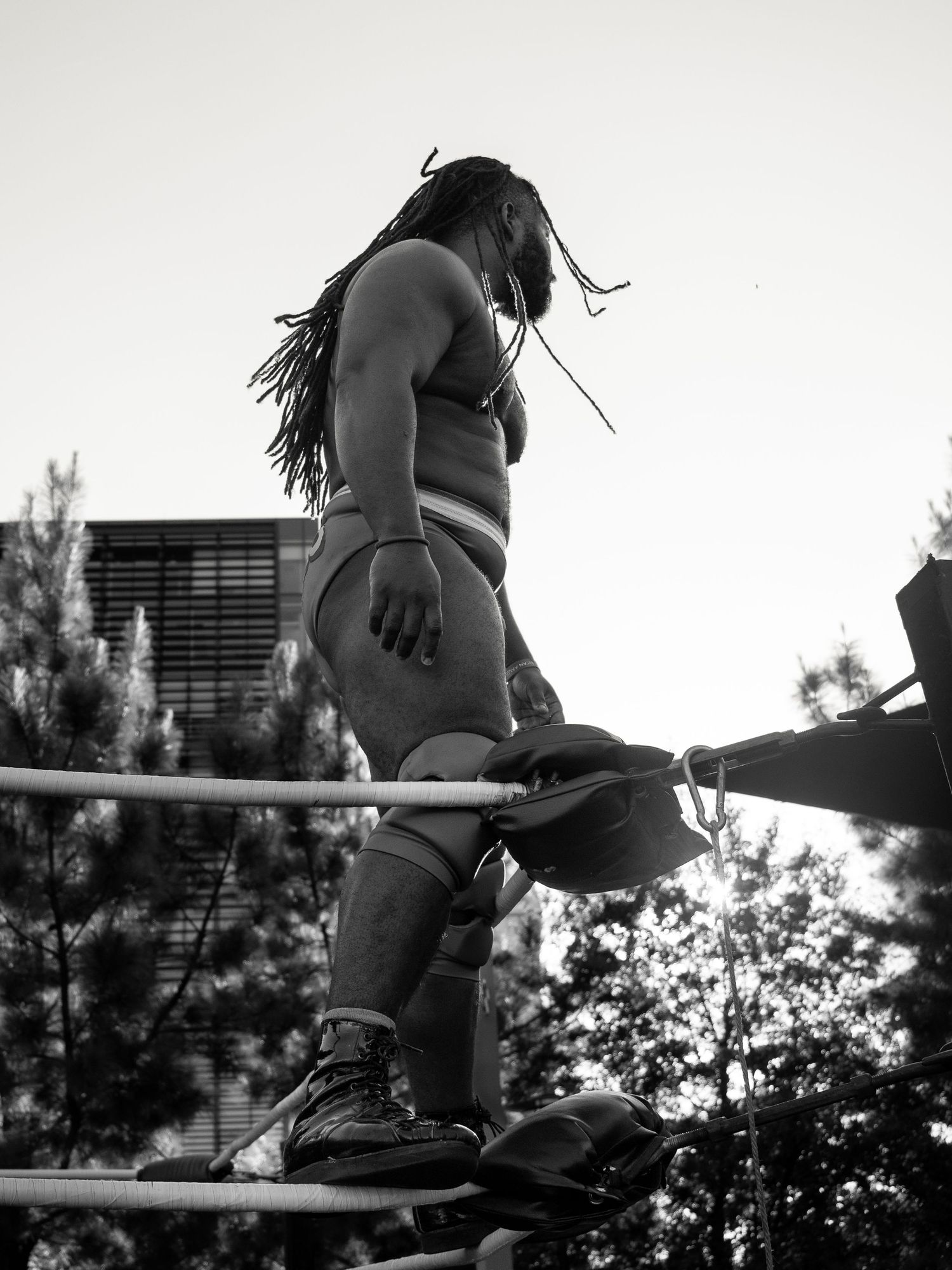 Black and white photo of a dreadlocked wrestler standing on ring ropes outdoors with trees in background.