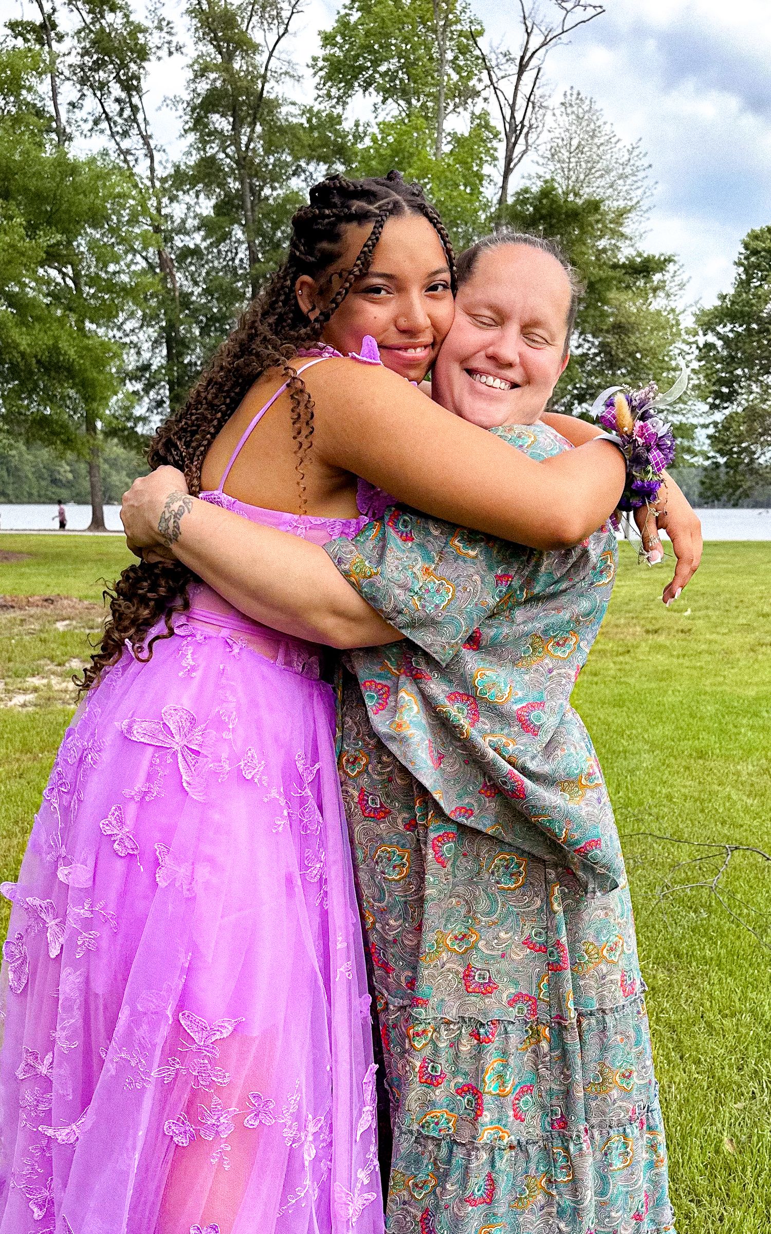 Two young women in formal dresses share a warm hug outdoors, one in a purple gown and one in a floral dress.