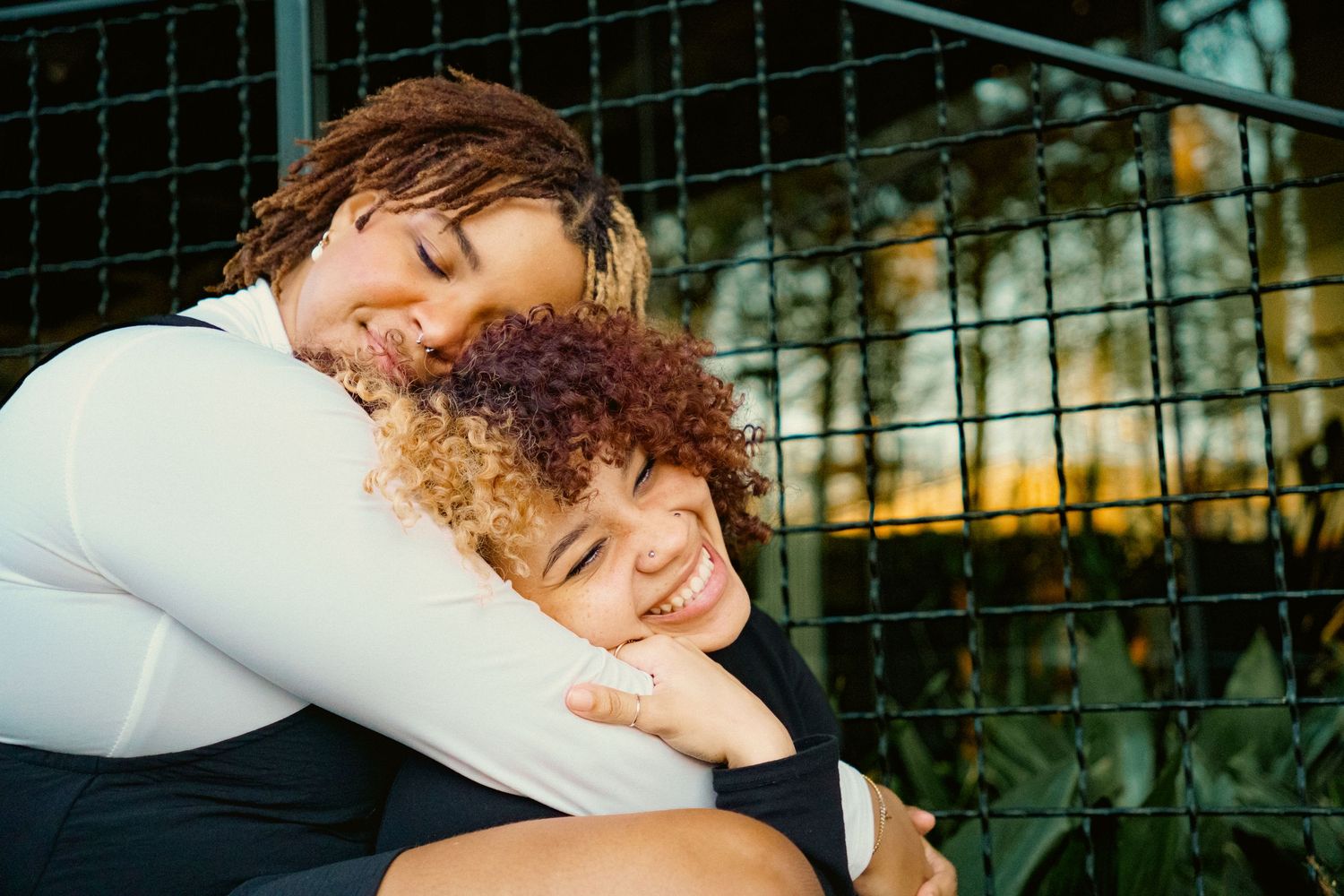 Two young women sharing a warm embrace outdoors, smiling joyfully near a metal fence at sunset.