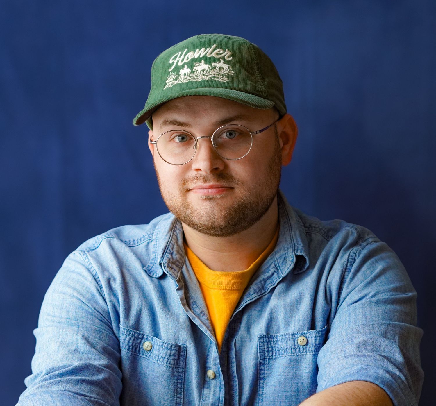 Young man wearing green cap and glasses with denim shirt poses for professional headshot against blue background.