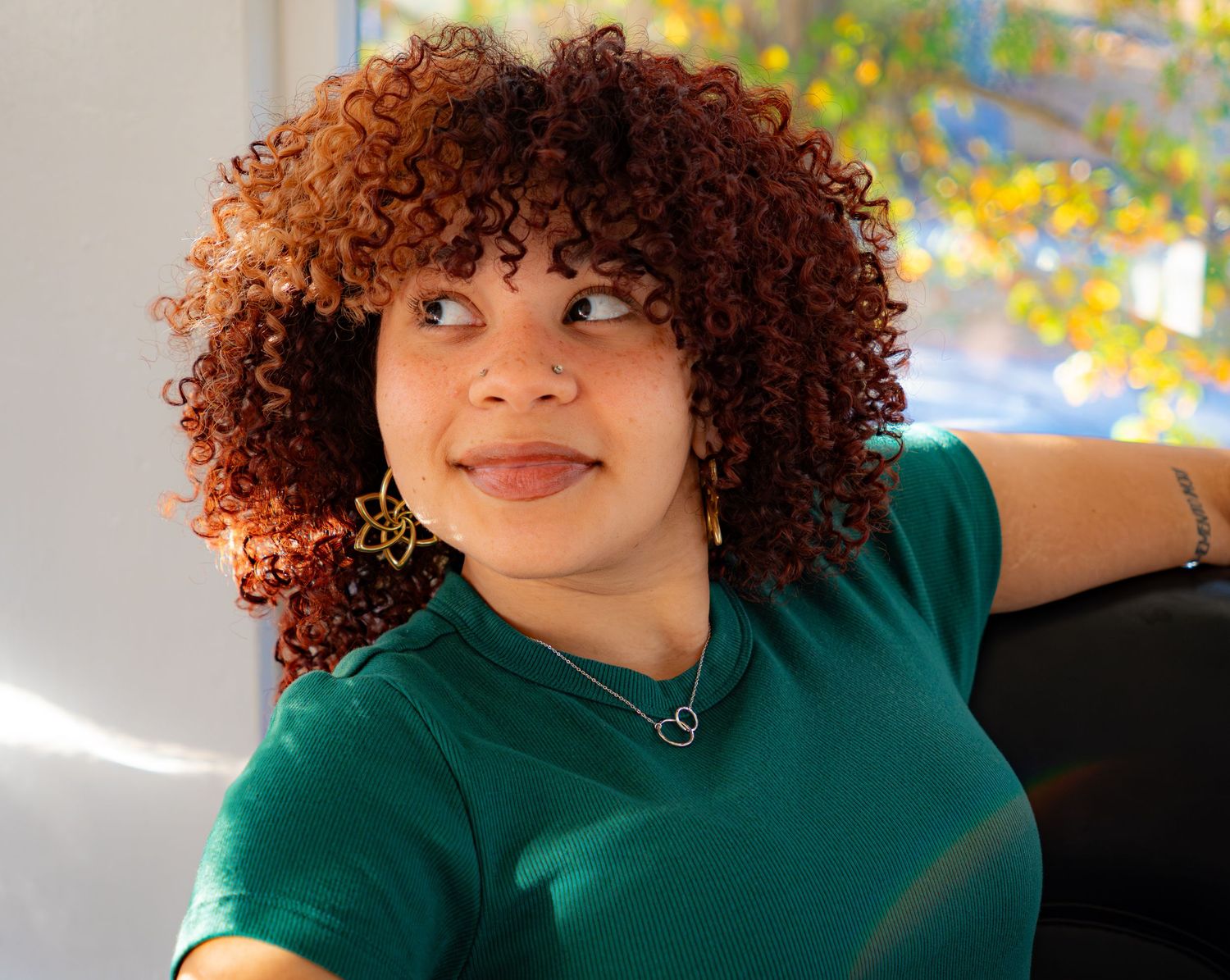 Young woman with vibrant curly auburn hair wearing a teal shirt and gold earrings, smiling indoors.