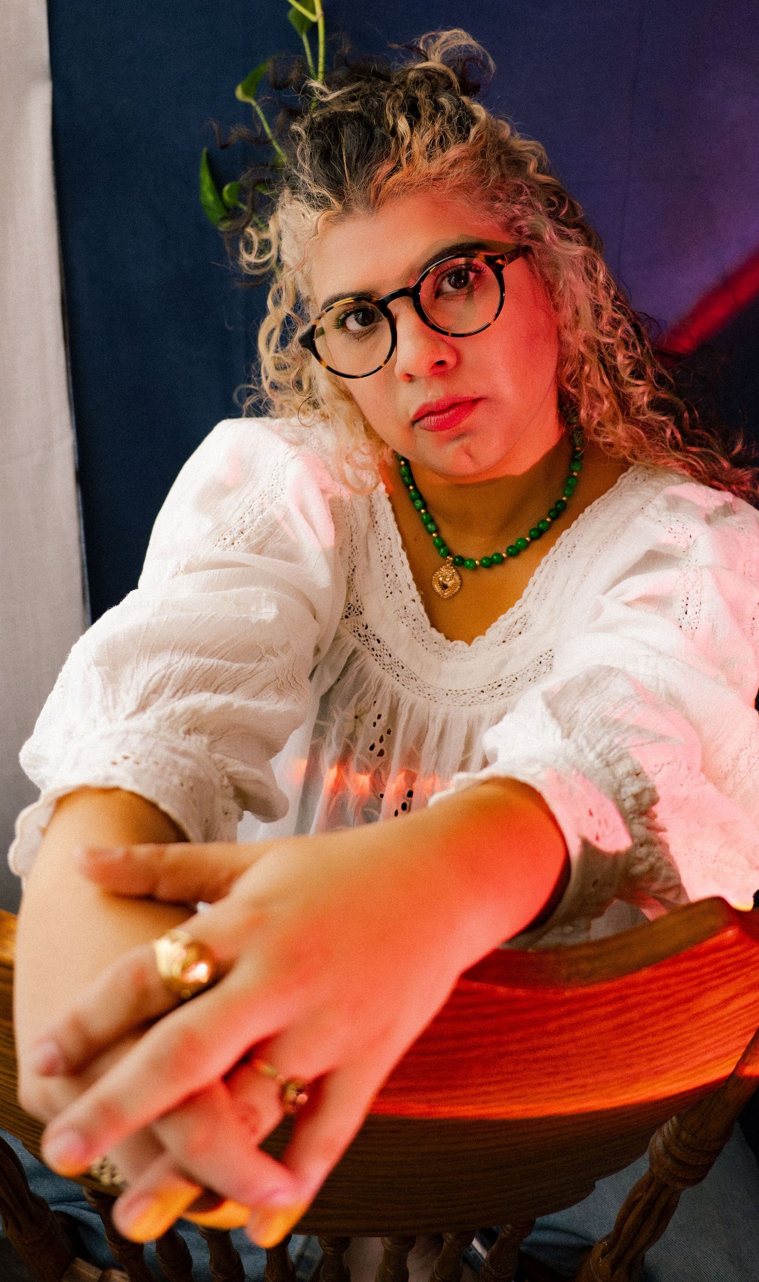 Young woman with curly hair, glasses, and green necklace posing in a white blouse.