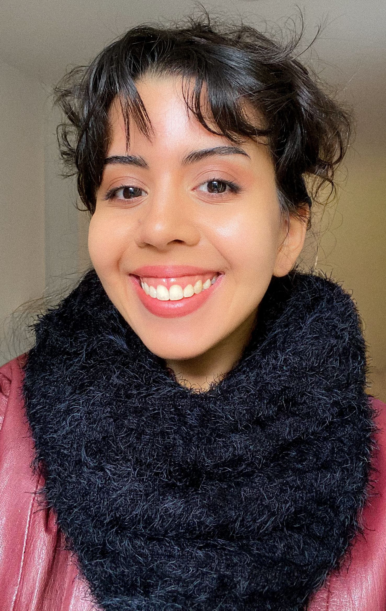 Young woman smiling warmly wearing black knit cowl scarf and burgundy jacket indoors.