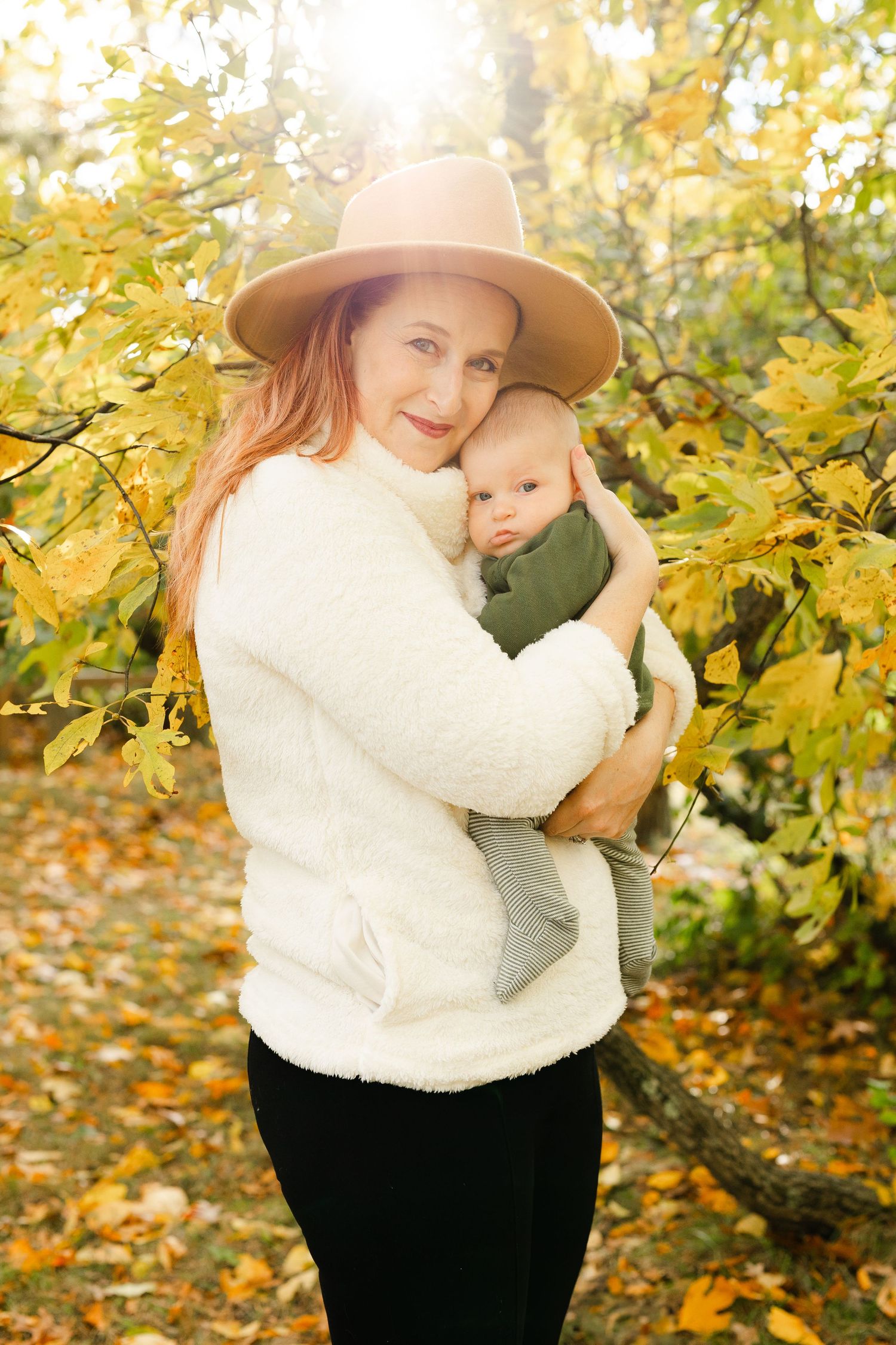 Red-haired mother in a wide-brim hat and white sweater cuddles her baby outdoors in autumn foliage.