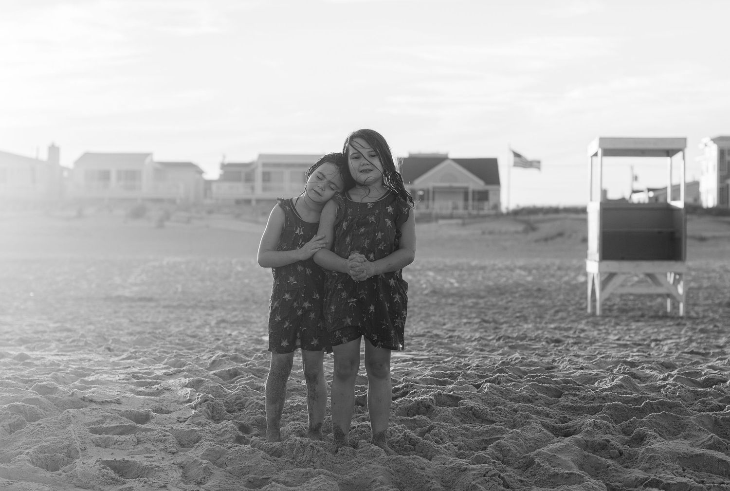 Two young girls hugging and smiling on a sandy beach in black and white photo with houses in background.