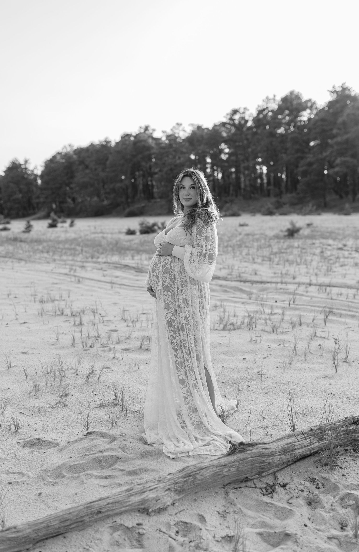 Black and white maternity photo of a woman in a flowing lace gown standing on a sandy beach near driftwood.
