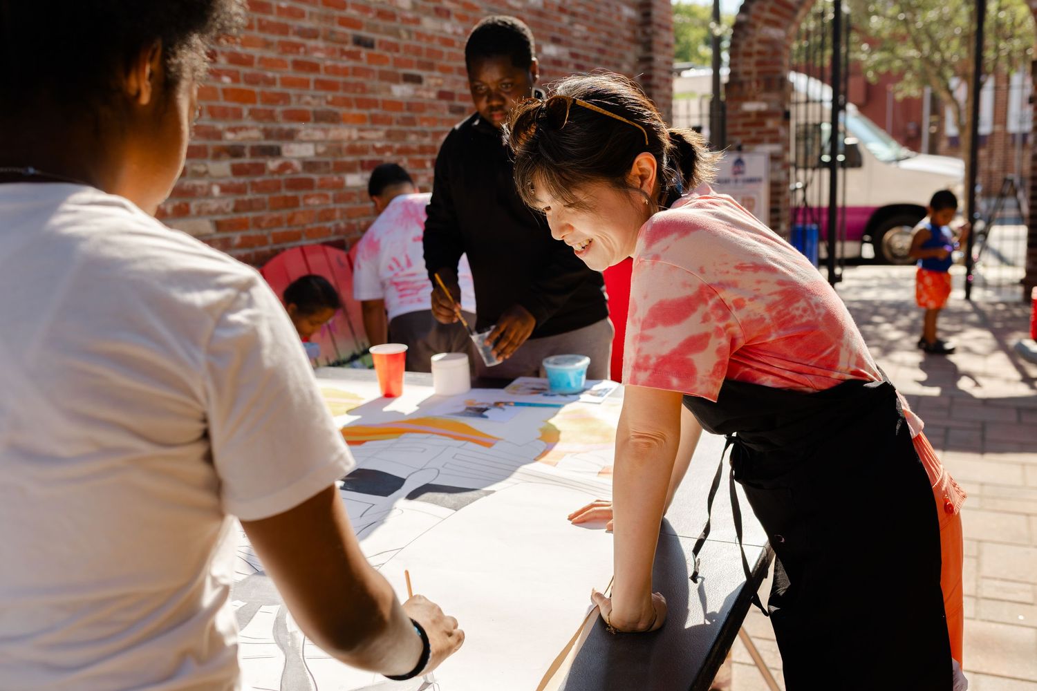 People collaborating on an outdoor art project at a table near a brick wall on a sunny day.