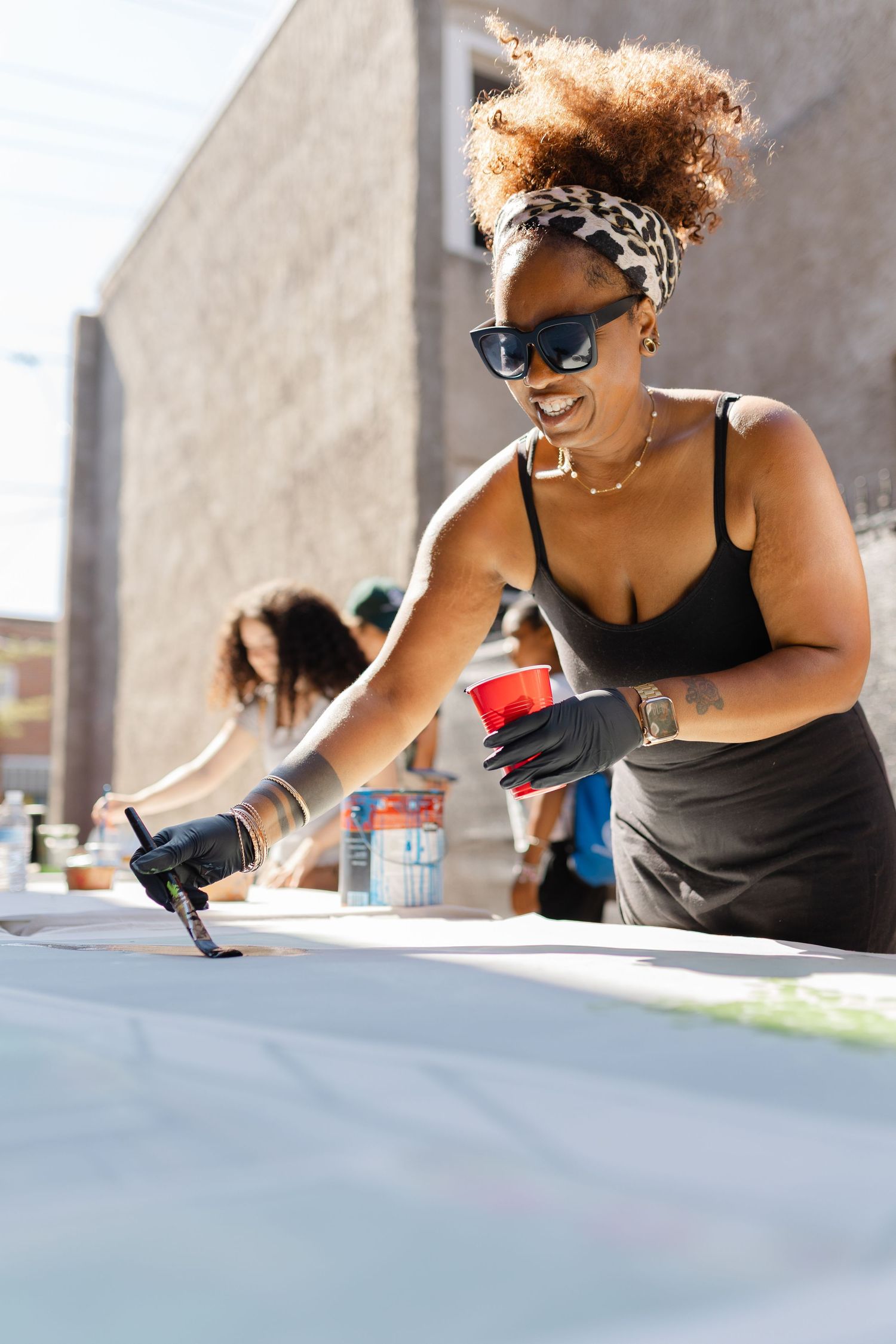 Smiling woman in sunglasses painting large white canvas outdoors on sunny day.
