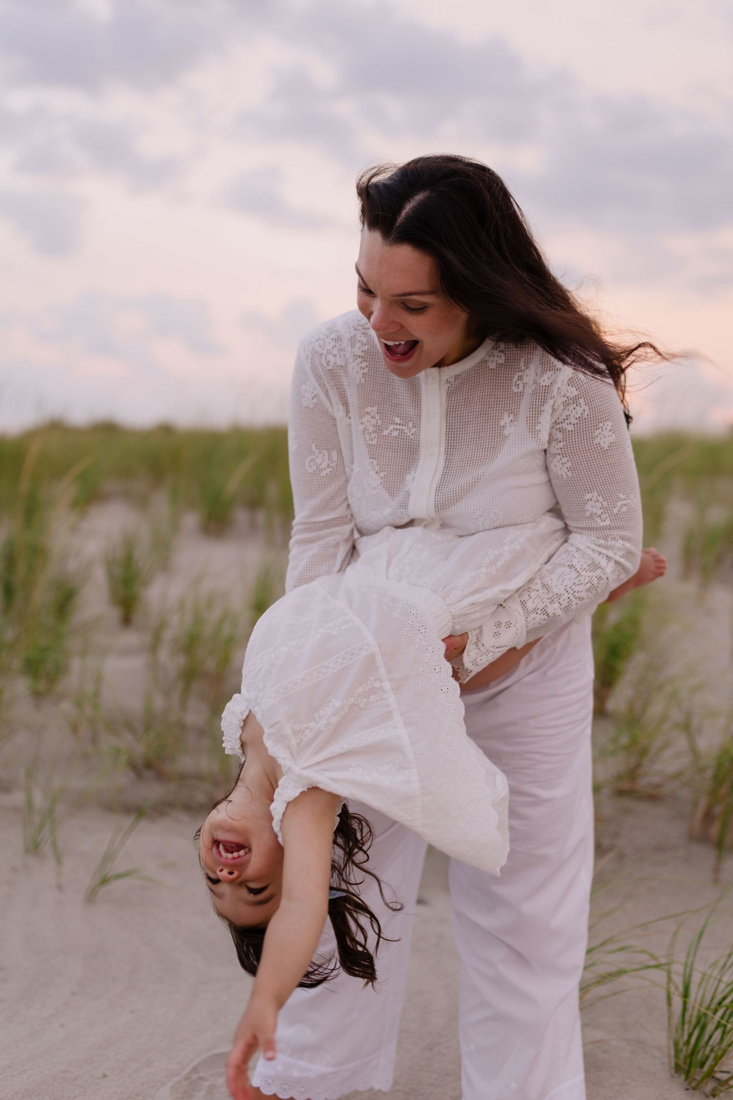 A laughing woman in white lace dress playfully holds a child upside down on a sandy beach at dusk.