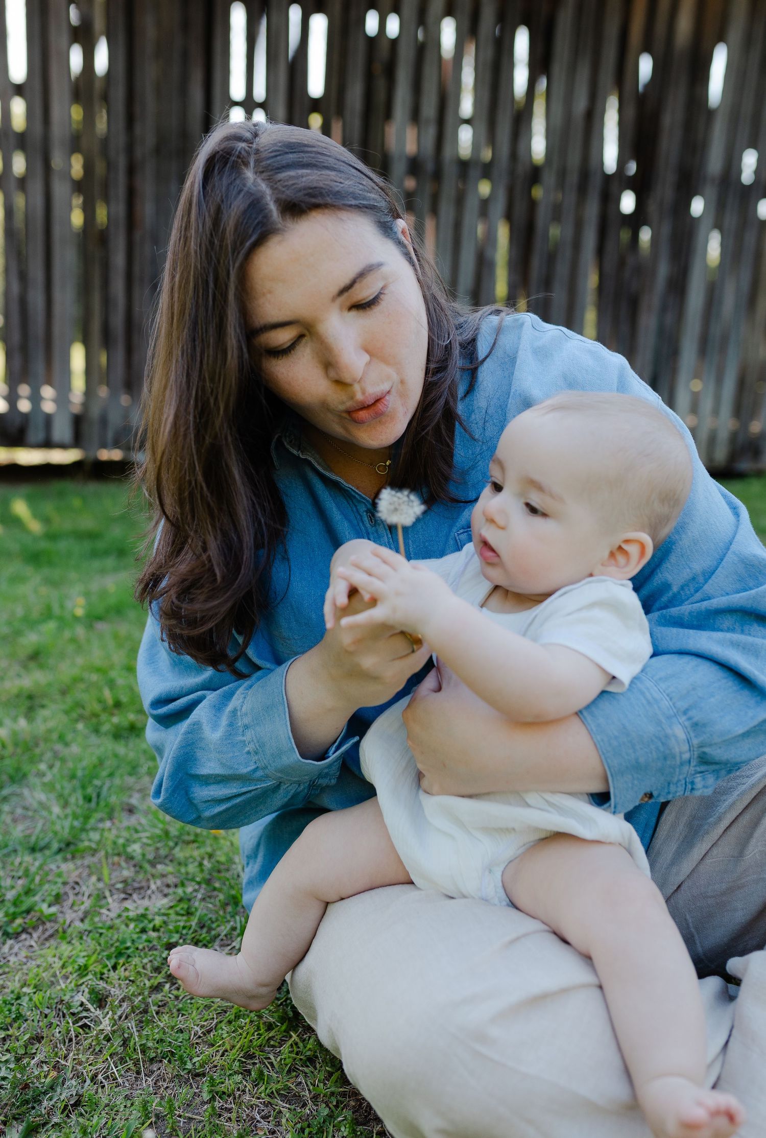 Mother sitting on grass blowing dandelion while curious baby reaches for it.