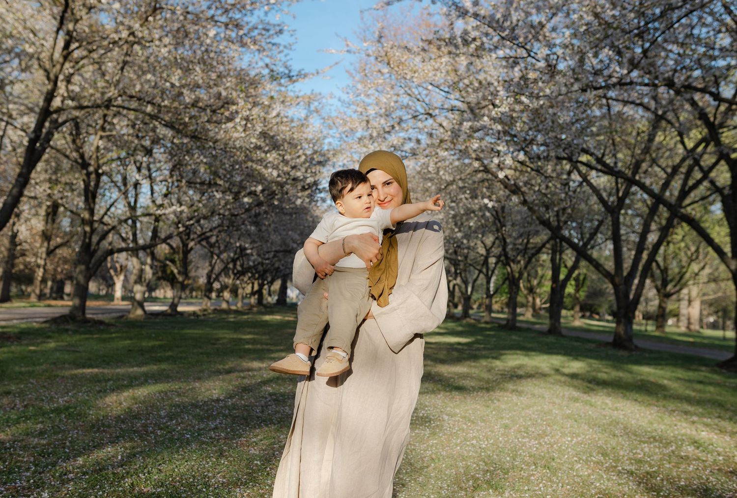 A smiling mother in a hijab holds her baby while standing among blooming white trees in a sunny park.