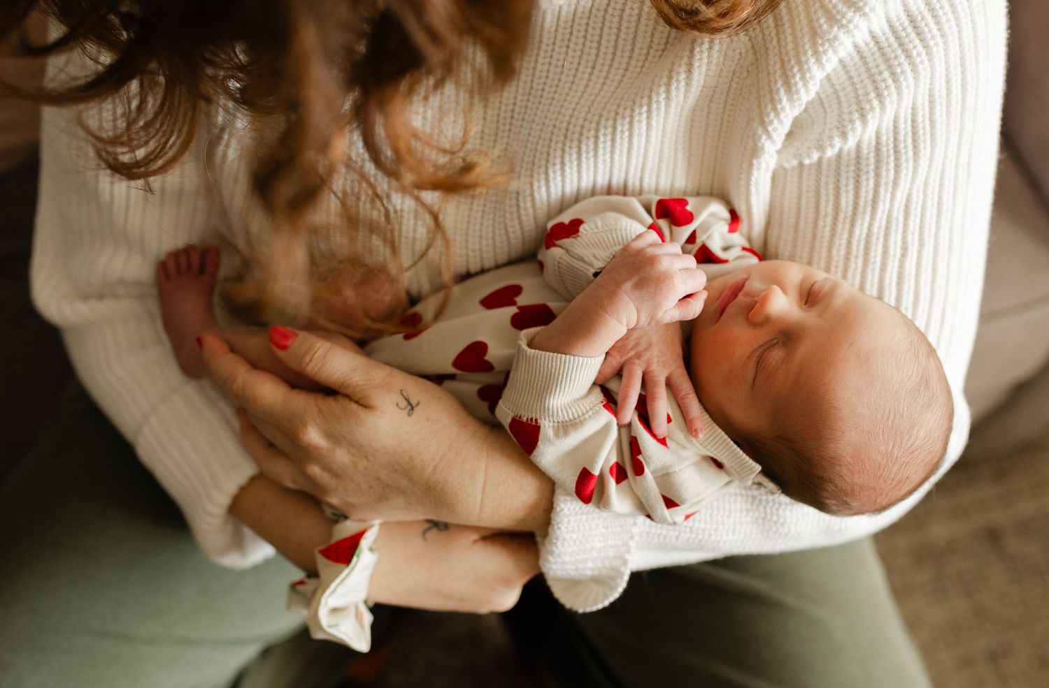 A mother in a white sweater cradles her sleeping newborn dressed in a floral outfit.