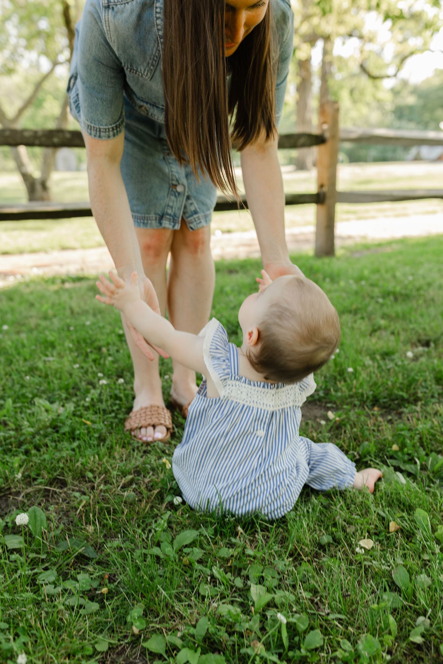 Mother helping baby sit up on green grass lawn in sunny outdoor park setting.