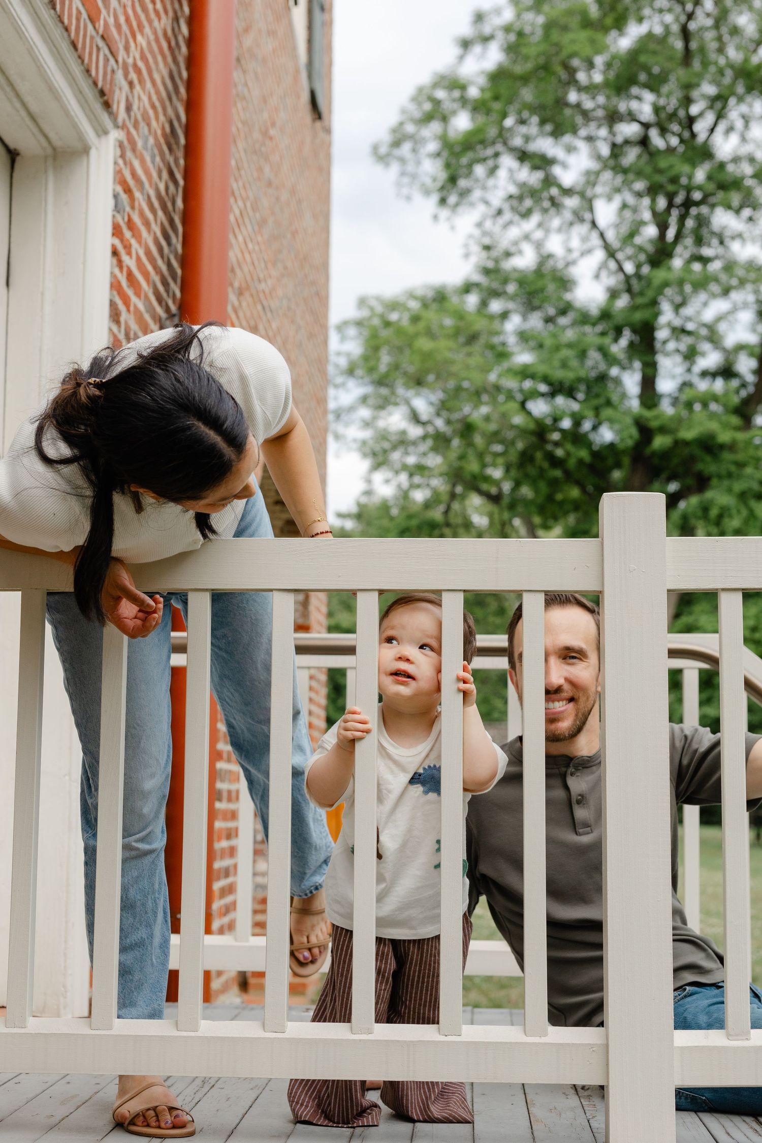 A young family with a toddler enjoying time on a white porch railing outside a brick home.