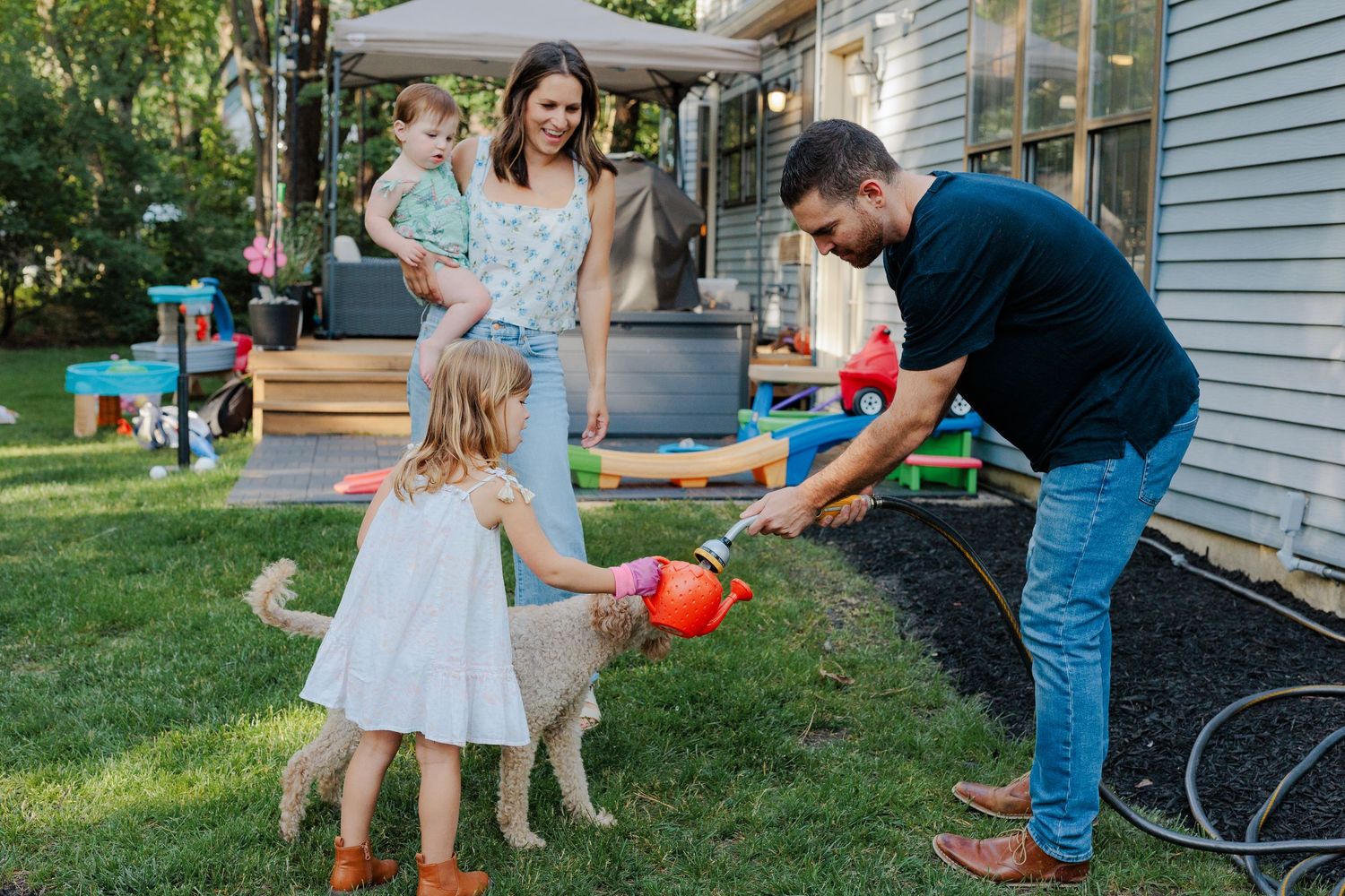 Family playing with a garden hose in a sunny backyard, parents and two young children enjoying outdoor water play.
