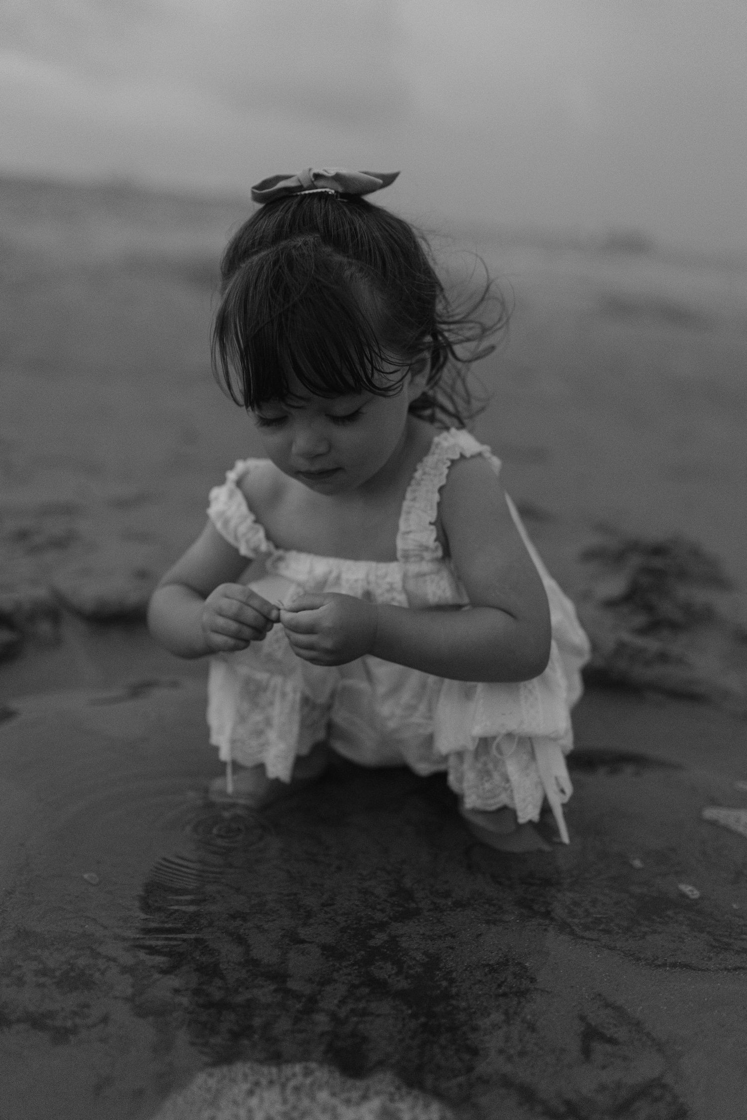 A young girl in a white dress plays in shallow beach water, looking down at her hands in a black and white photo.