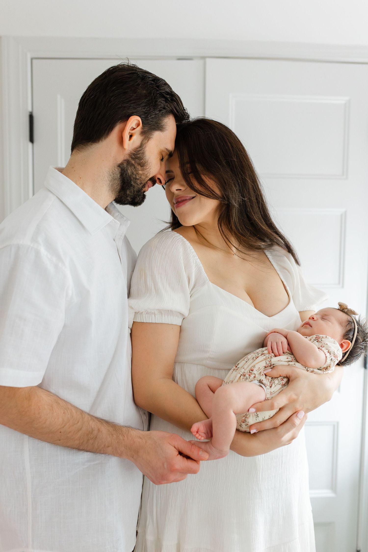 New parents in white outfits touching foreheads while mother holds sleeping newborn baby girl.