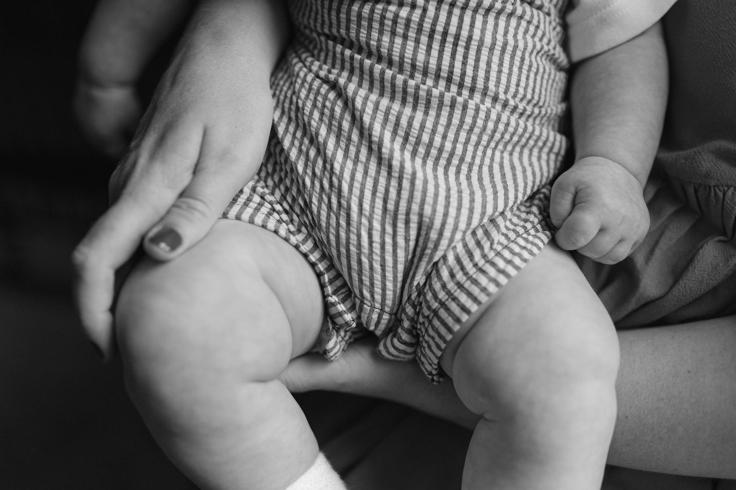 Close-up black and white photo of a baby in striped romper sitting on an adults lap.