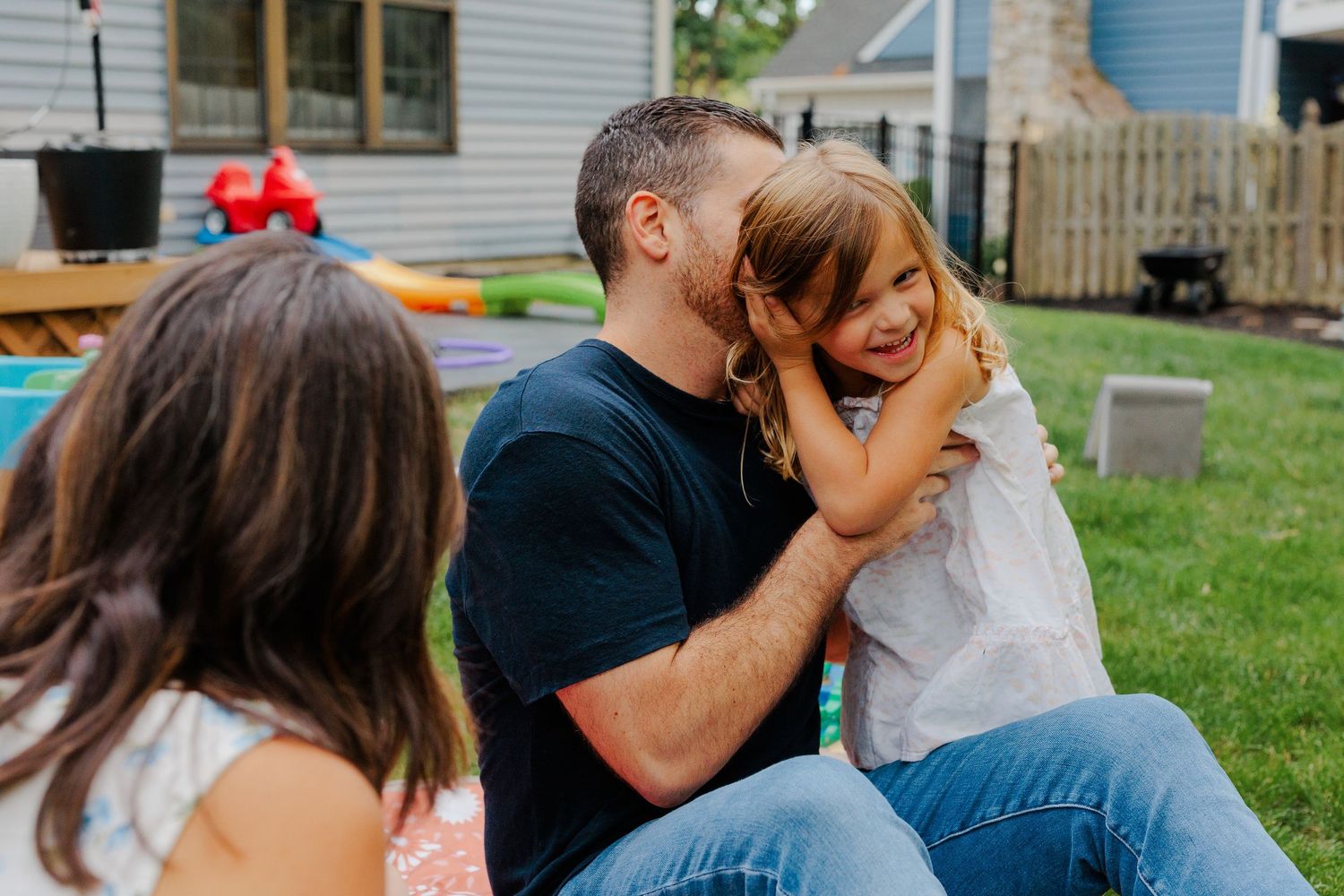 Father kissing smiling young daughter on the cheek while sitting outdoors in a backyard.