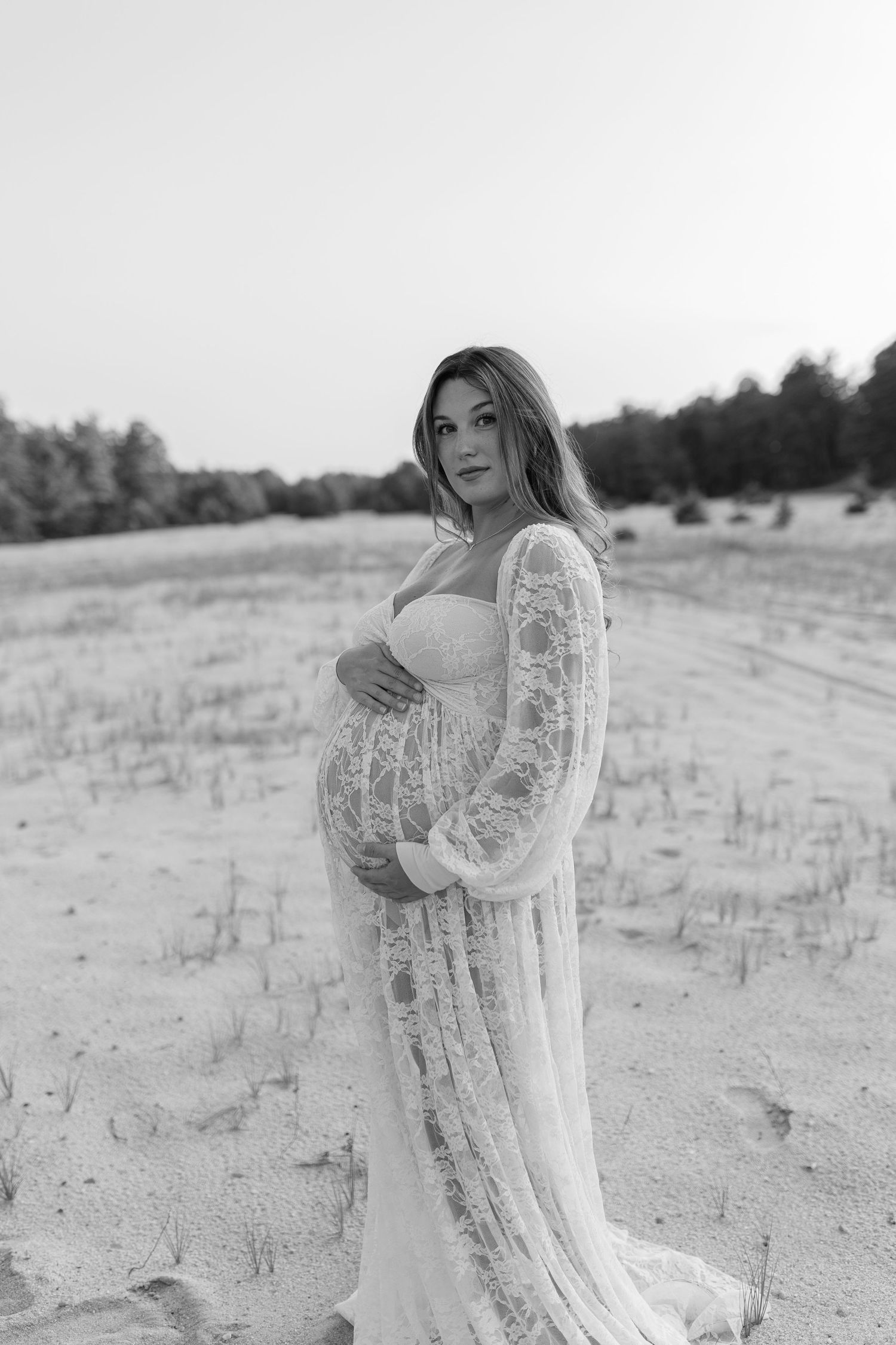 Expectant mother in flowing white lace dress cradles baby bump on sandy beach in black and white.