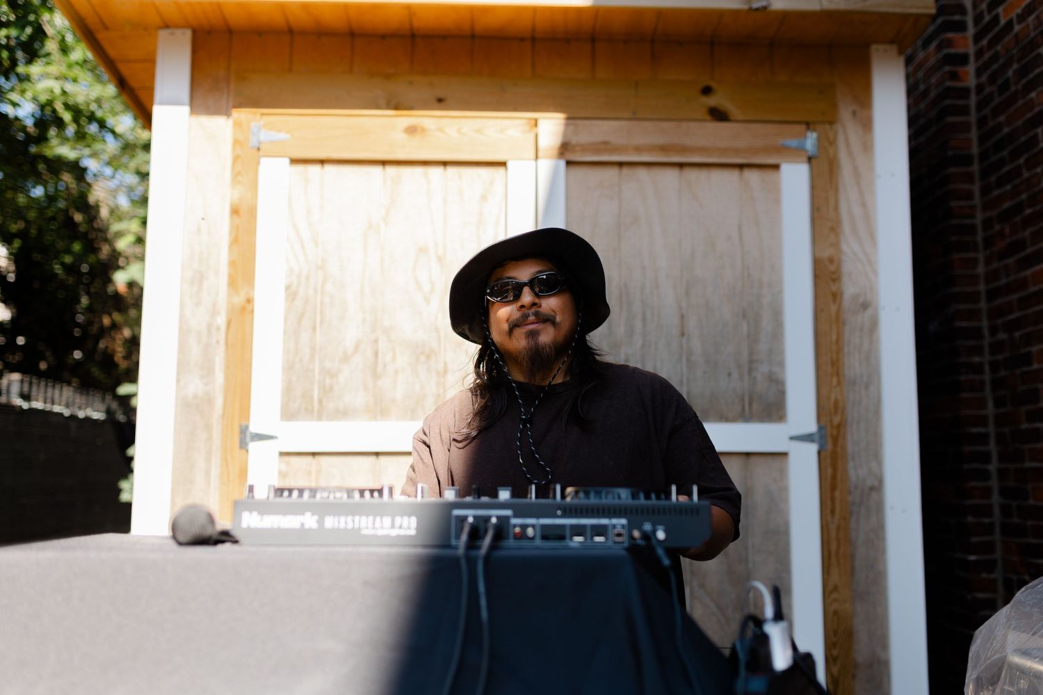 Man in black hat playing keyboard synthesizer outdoors near wooden structure on sunny day.