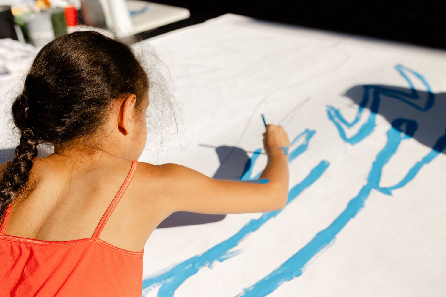 Young girl with braids in red top painting blue strokes on large white canvas in art class.