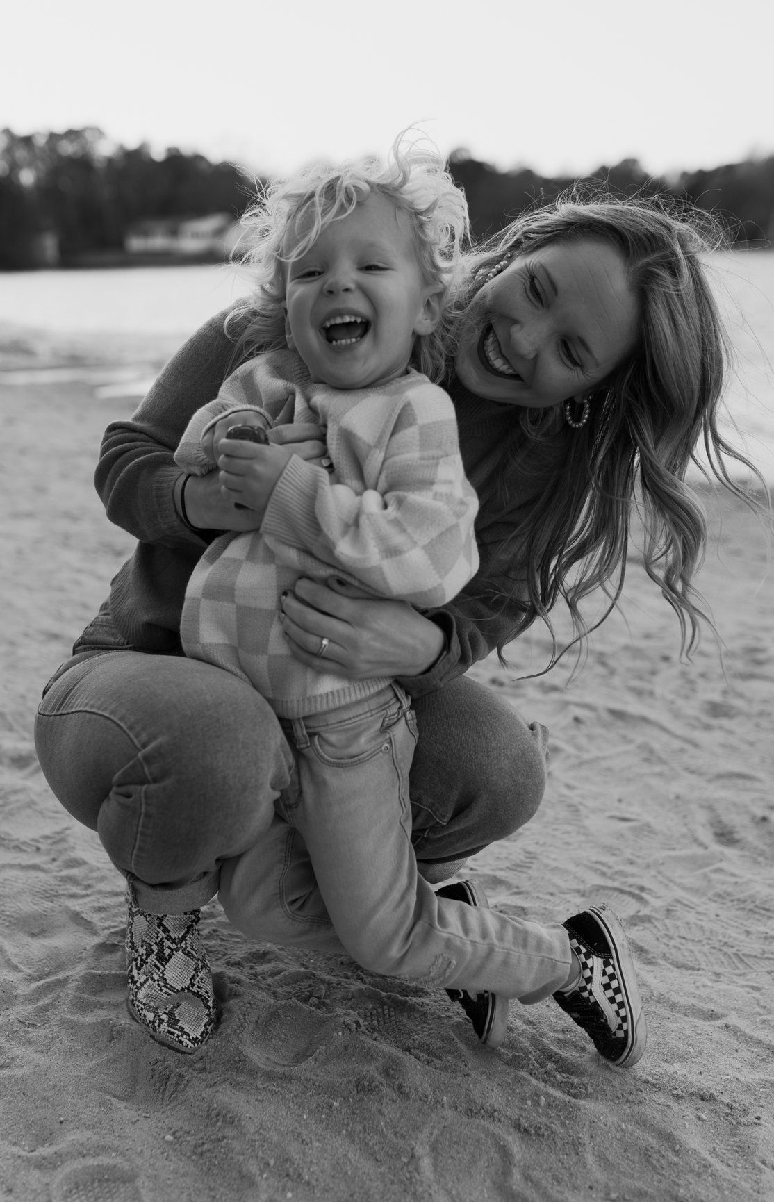 Black and white photo of a smiling mother hugging her laughing toddler on a sandy beach.