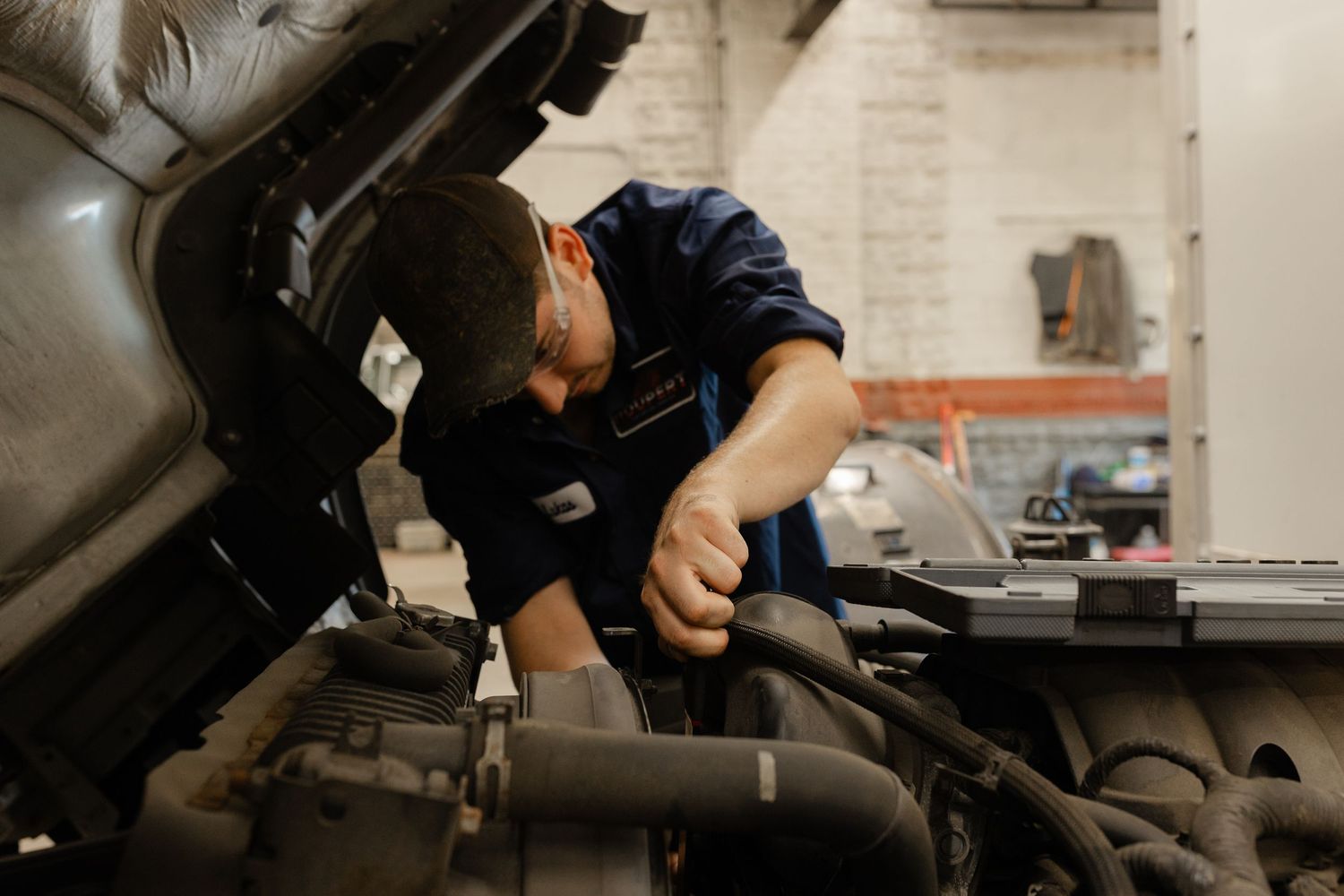 A mechanic in blue uniform leans over an open car engine, performing repairs in an auto shop.