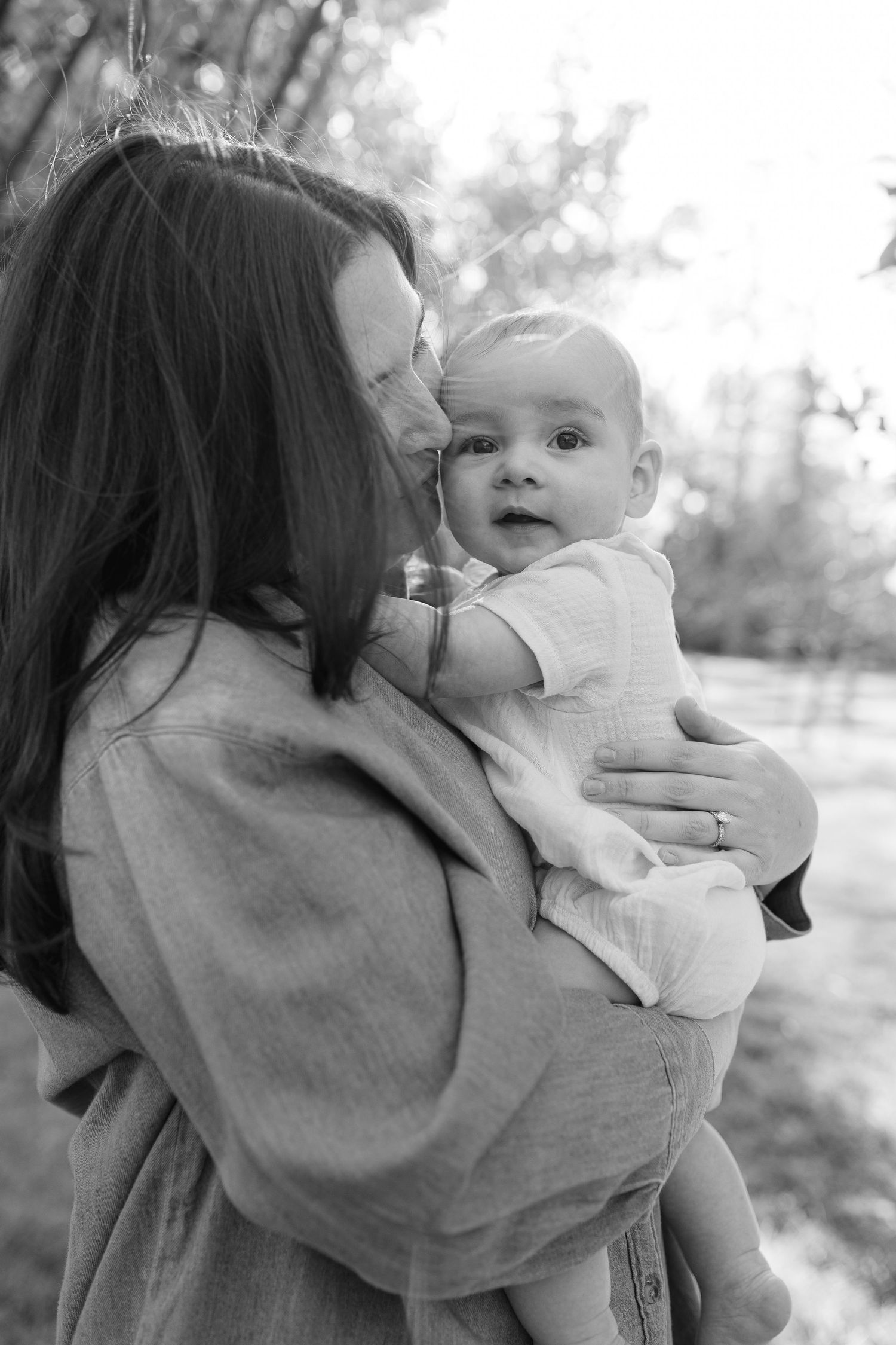 Black and white photo of a mother tenderly kissing her infant baby outdoors in a park setting.