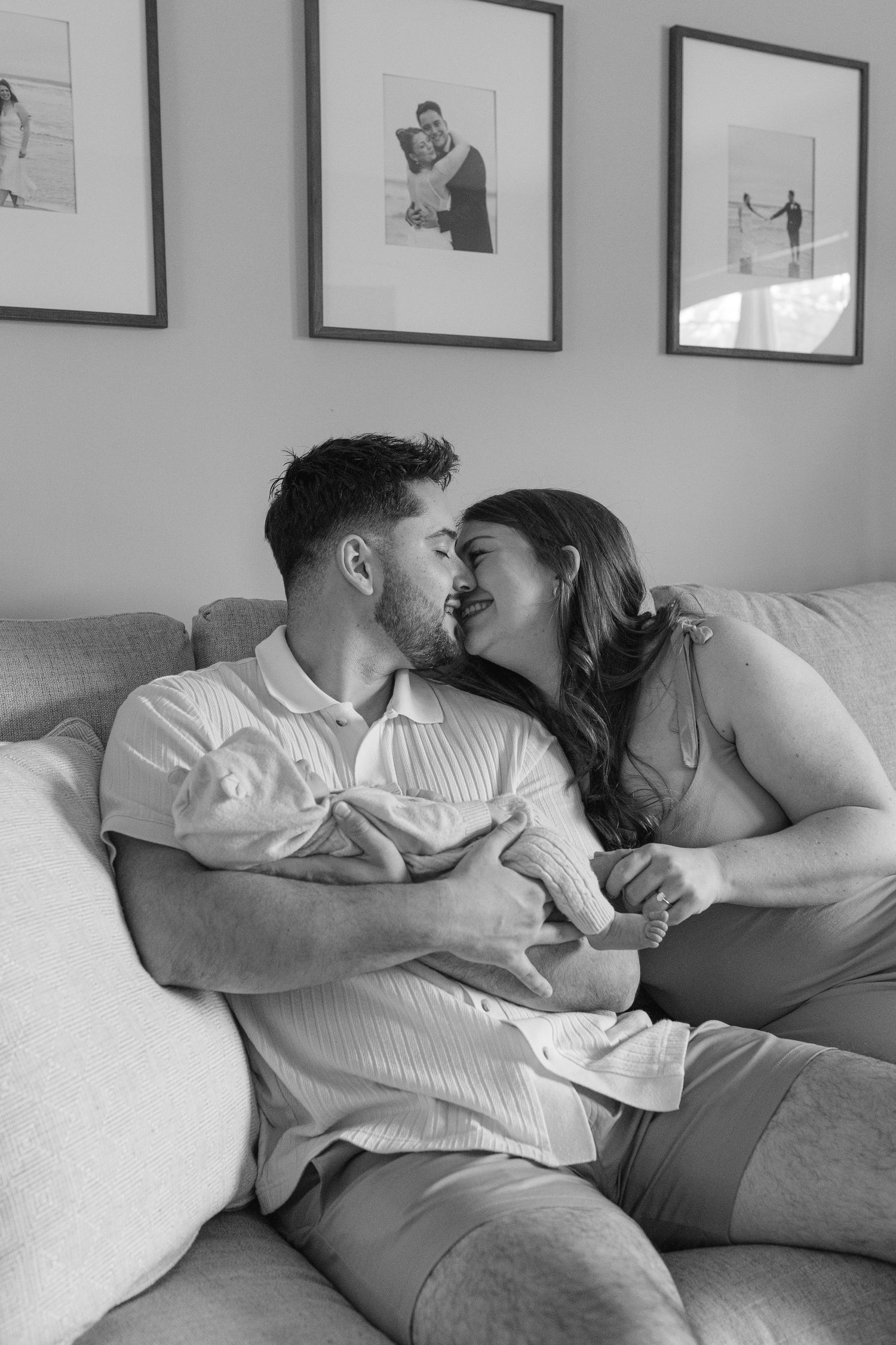 Black and white photo of couple kissing on couch while holding newborn baby together.