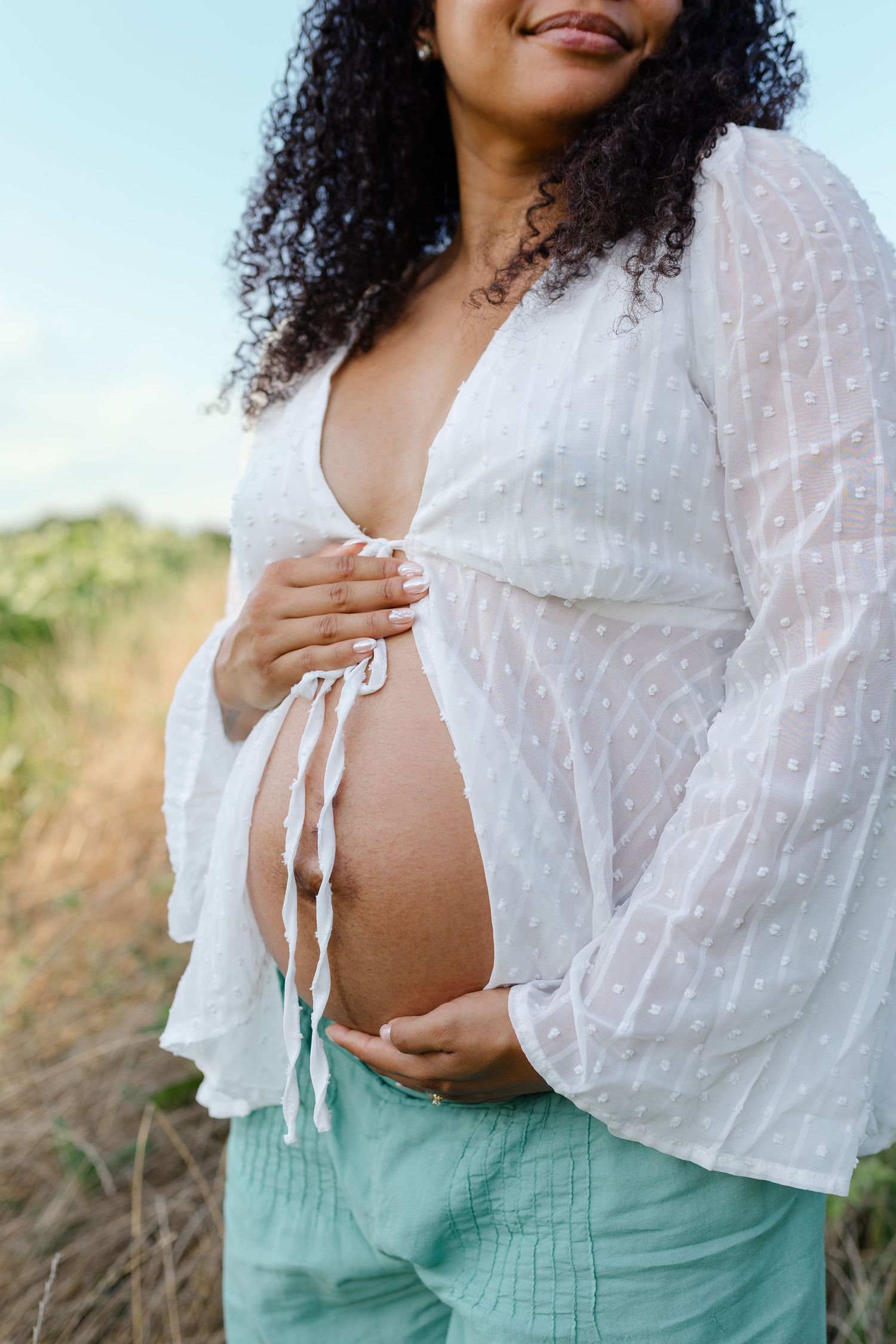 Smiling pregnant woman in sheer white top and mint pants gently holding her exposed belly outside.
