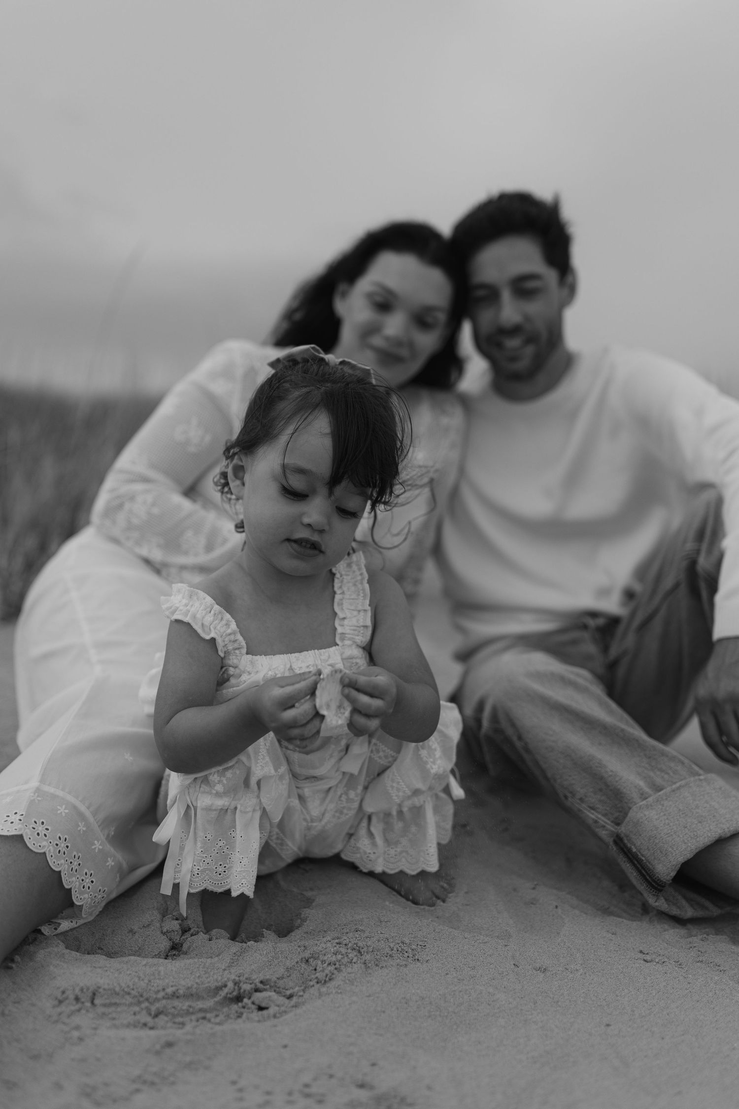 Black and white photo of a young family sitting on beach sand, parents smiling as toddler girl plays.
