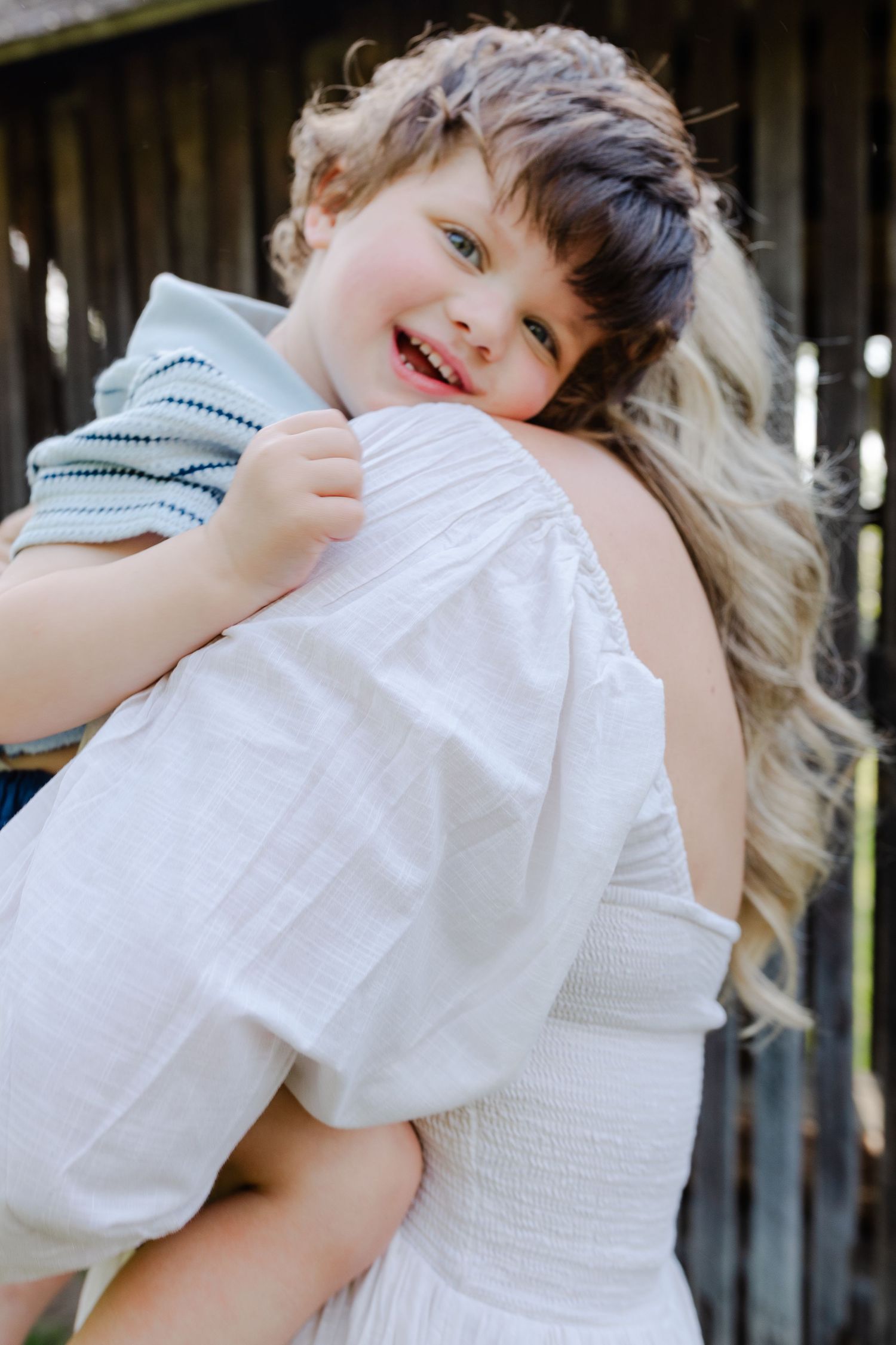 Smiling young boy clinging to his blonde mother wearing a white dress near a wooden structure.