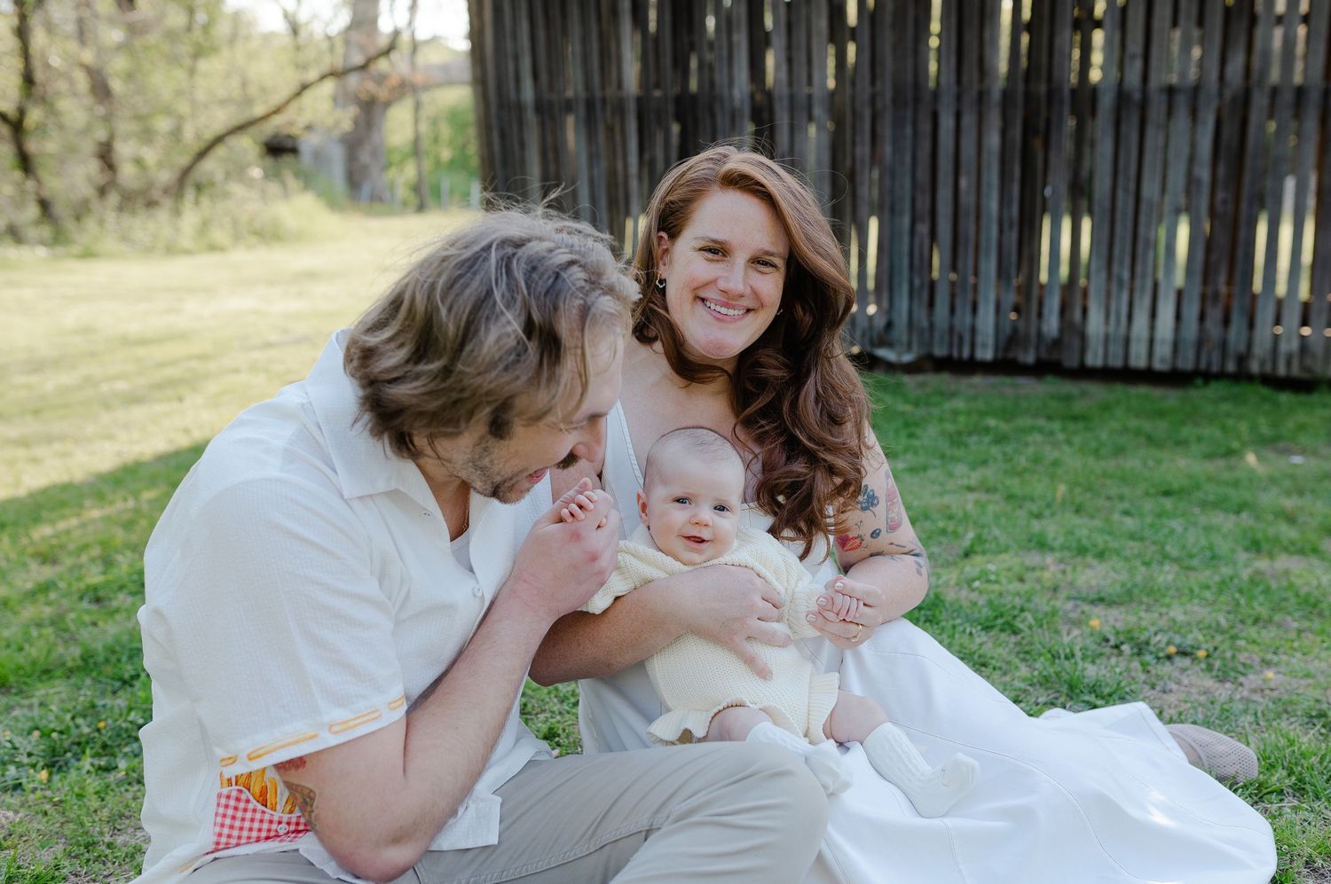 A happy family sits on grass outdoors, with a smiling mother holding a baby while a man leans in affectionately.