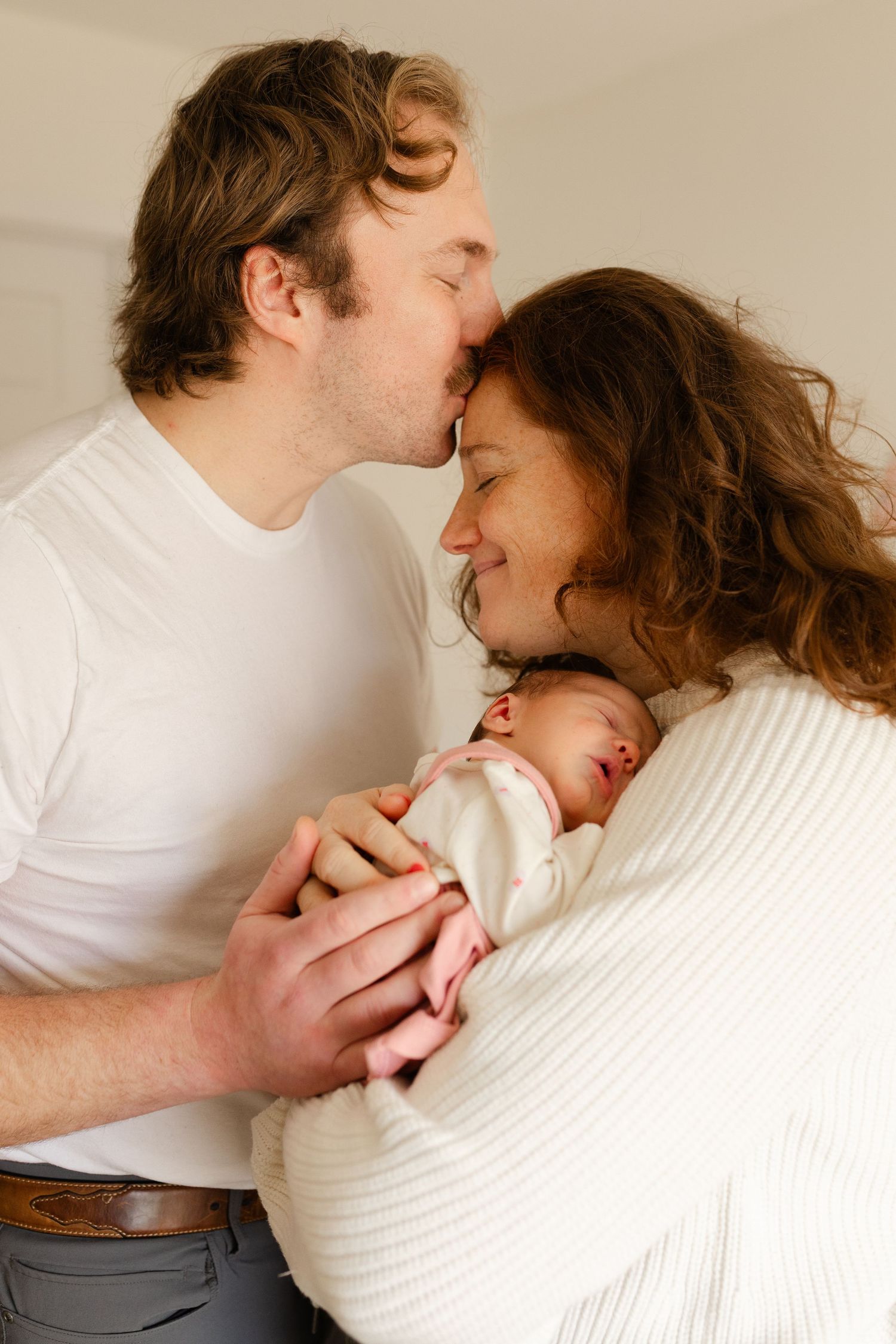 Happy couple sharing a kiss while cradling their newborn baby together in a bright room.