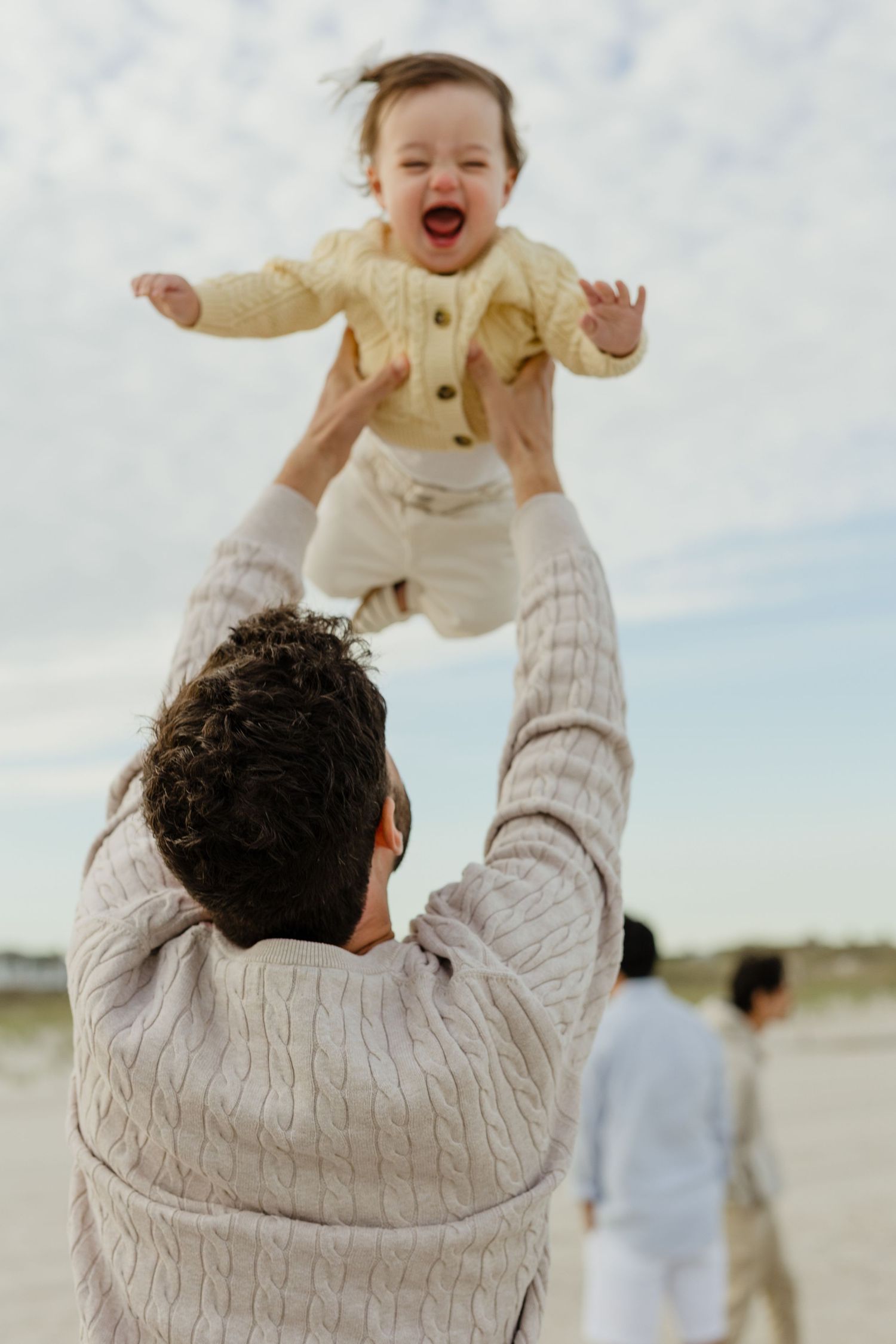 Joyful baby in yellow knit outfit being lifted high in the air on a beach.