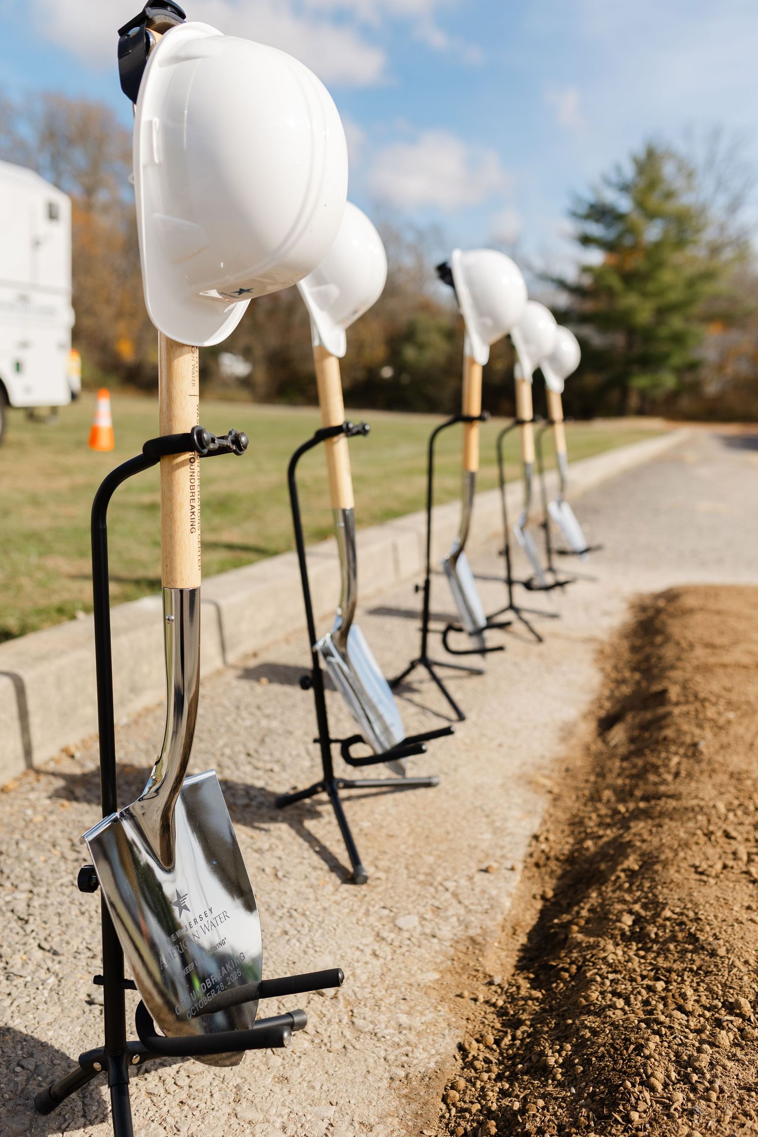 Row of white hard hats on posts along a dirt path during a groundbreaking ceremony.