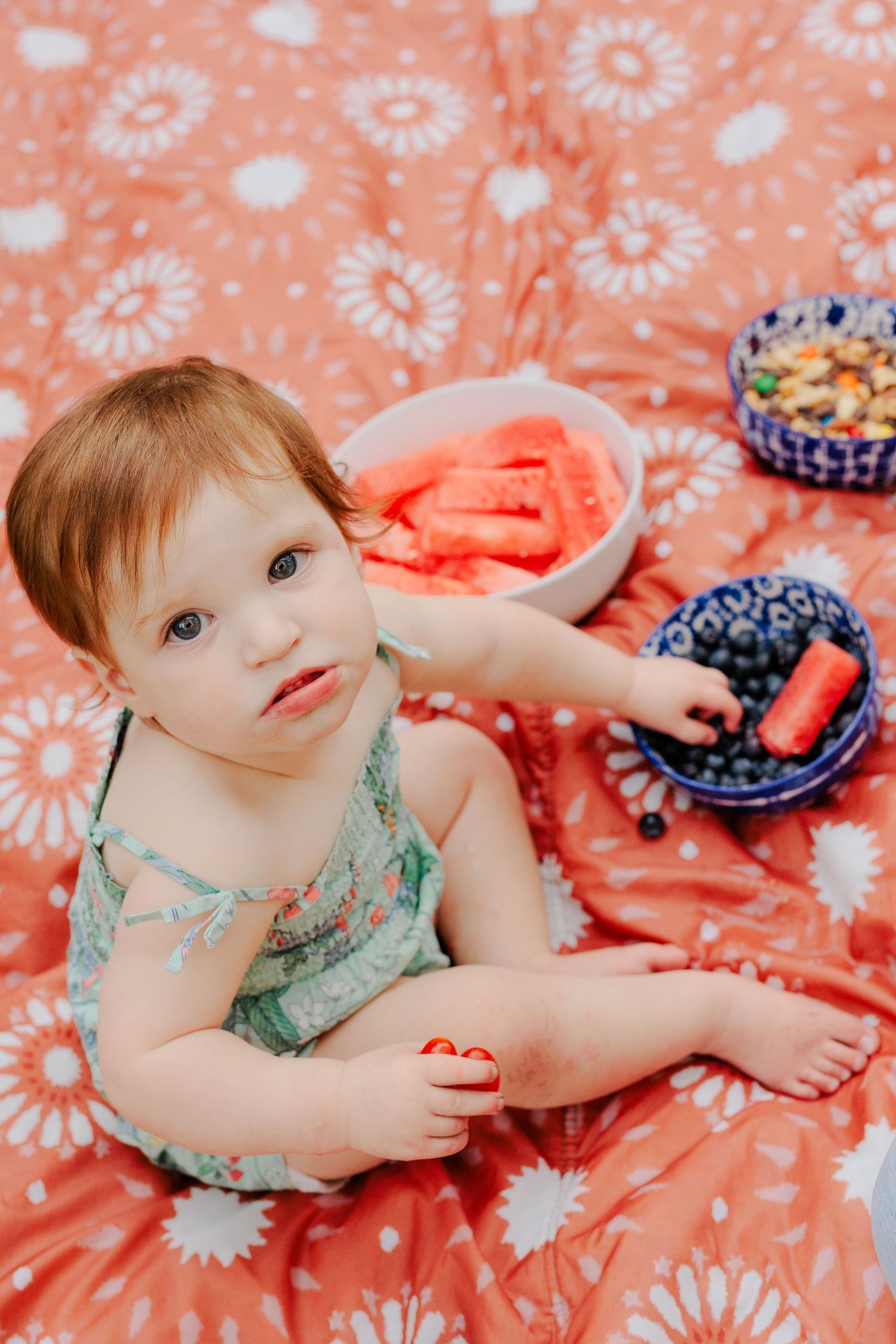 Baby girl sitting on red floral blanket eating fresh blueberries and watermelon slices at a summer picnic.