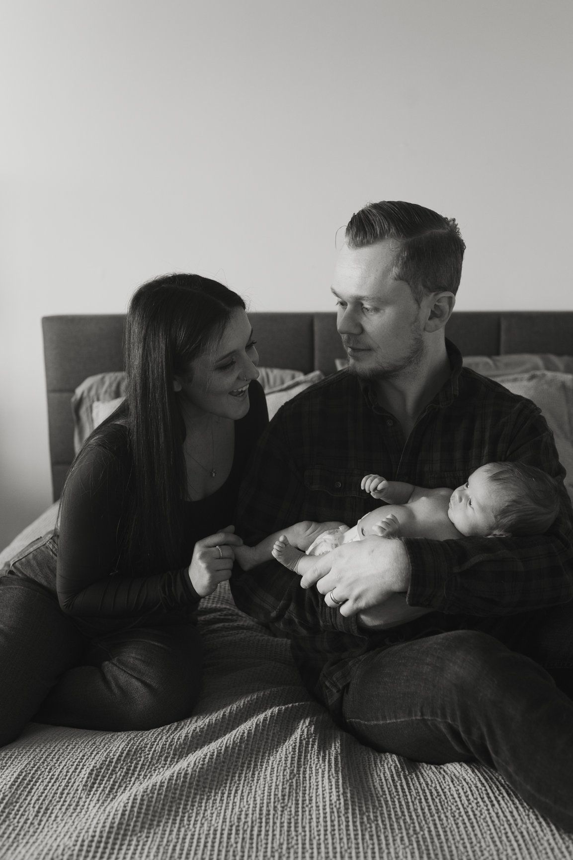 Young couple sitting on bed smiling at each other while holding their newborn baby in black and white.