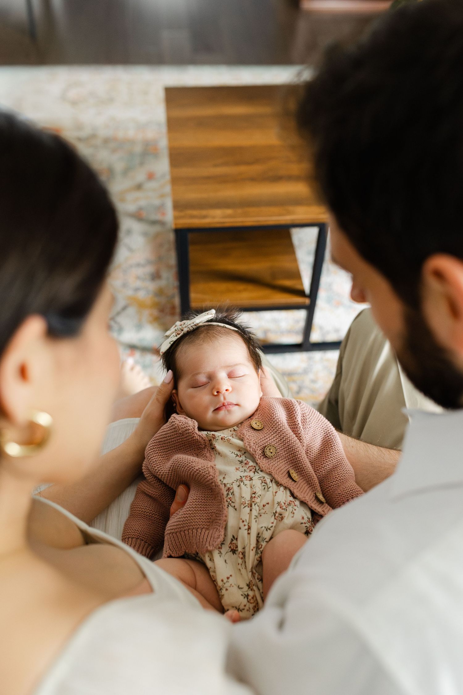 New parents looking down at their sleeping newborn baby girl in pink cardigan and floral headband.