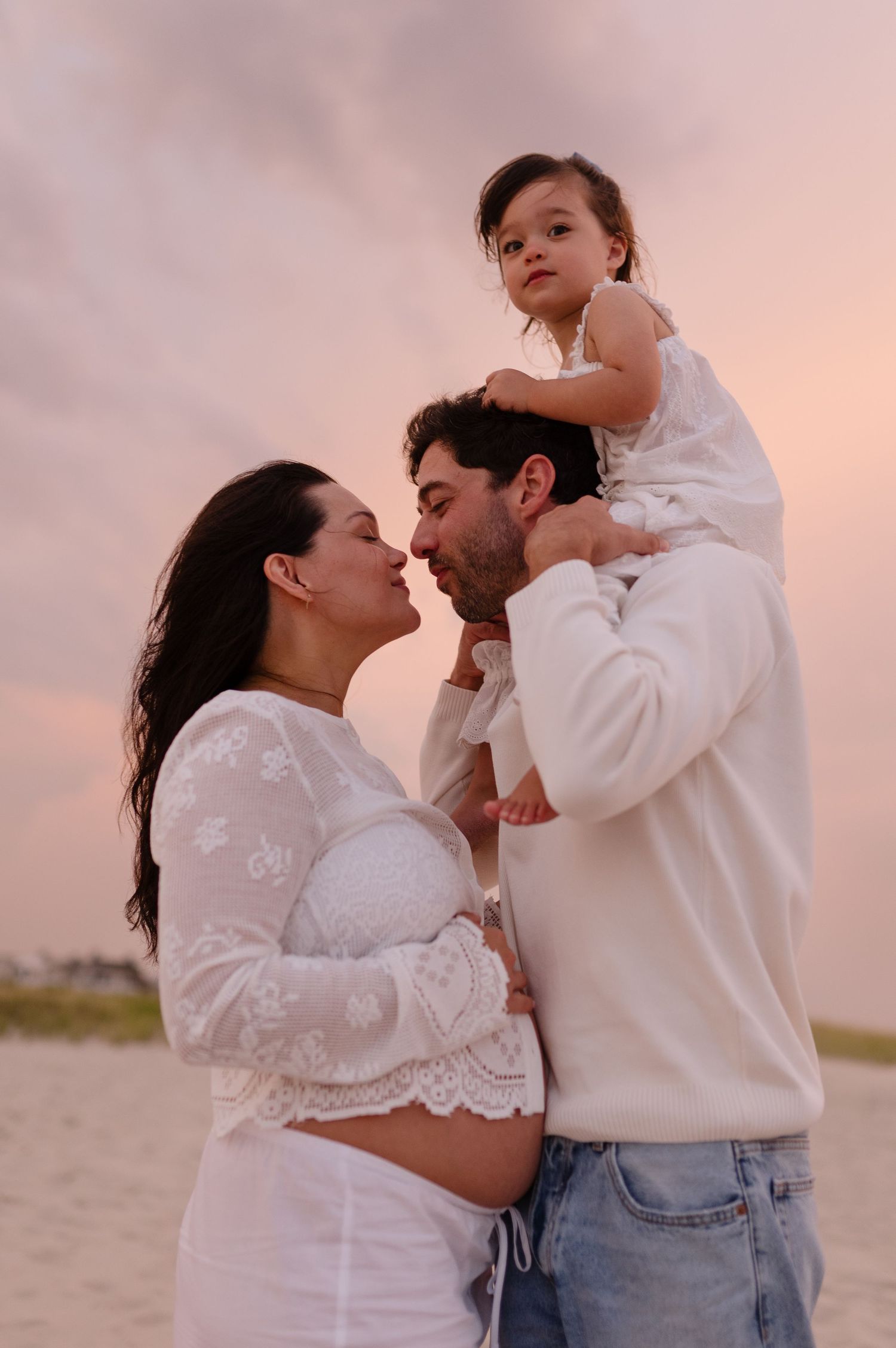 Pregnant woman and man sharing a kiss at sunset on a beach, with a toddler girl in white dress sitting on his shoulders.