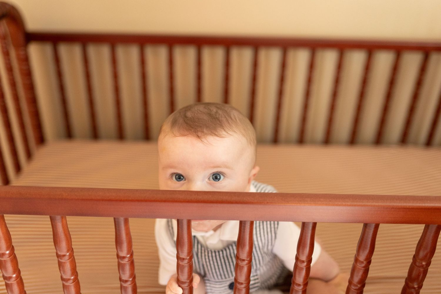 Curious blue-eyed baby peeking over the rail of a wooden crib.