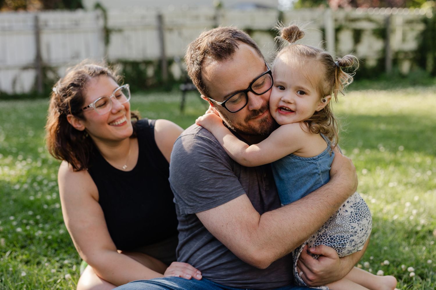 Happy family portrait of smiling parents cuddling their young toddler daughter while sitting on green grass.