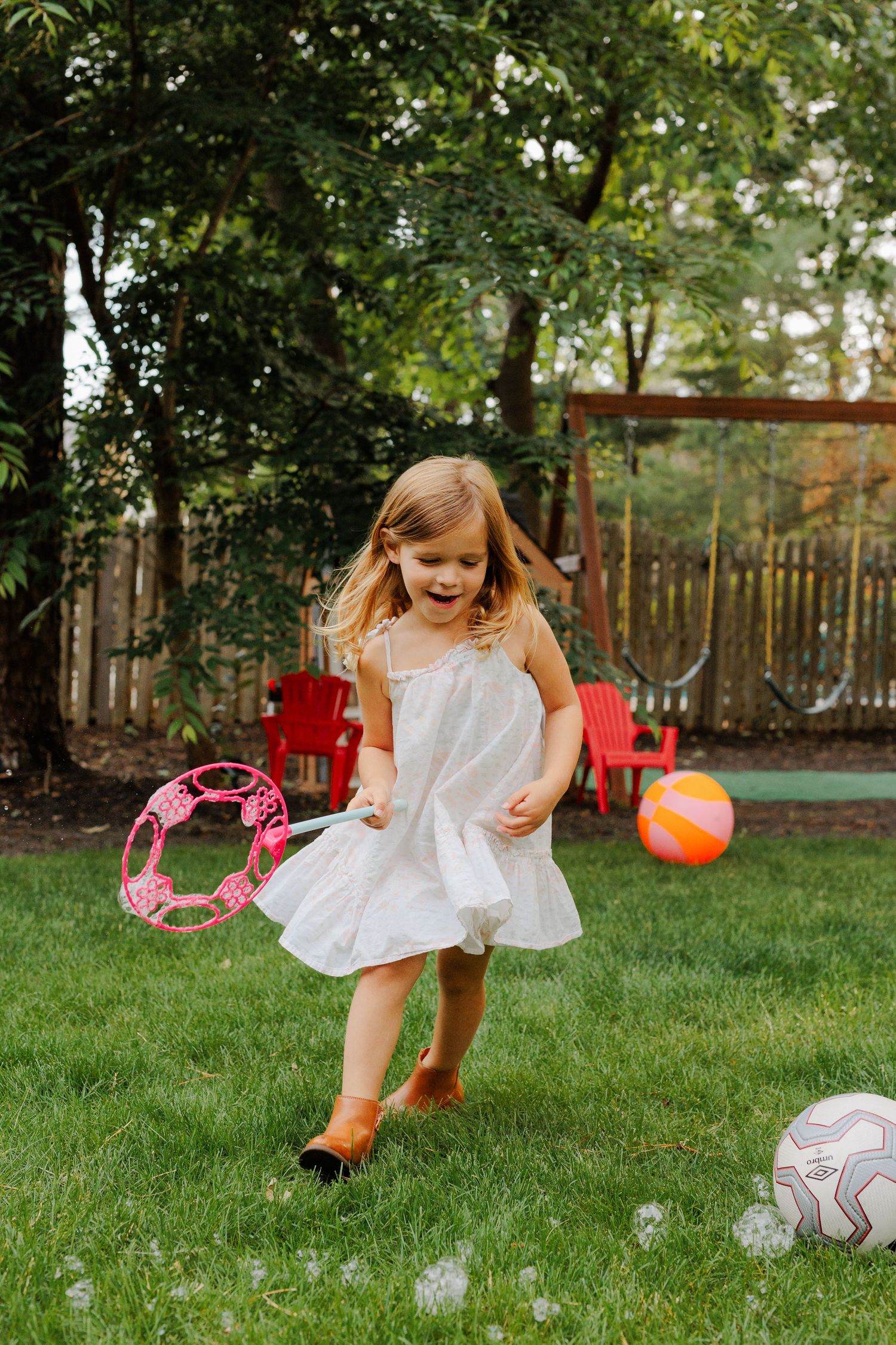 Happy little girl in white dress running on grass with pink bubble wand and colorful ball.