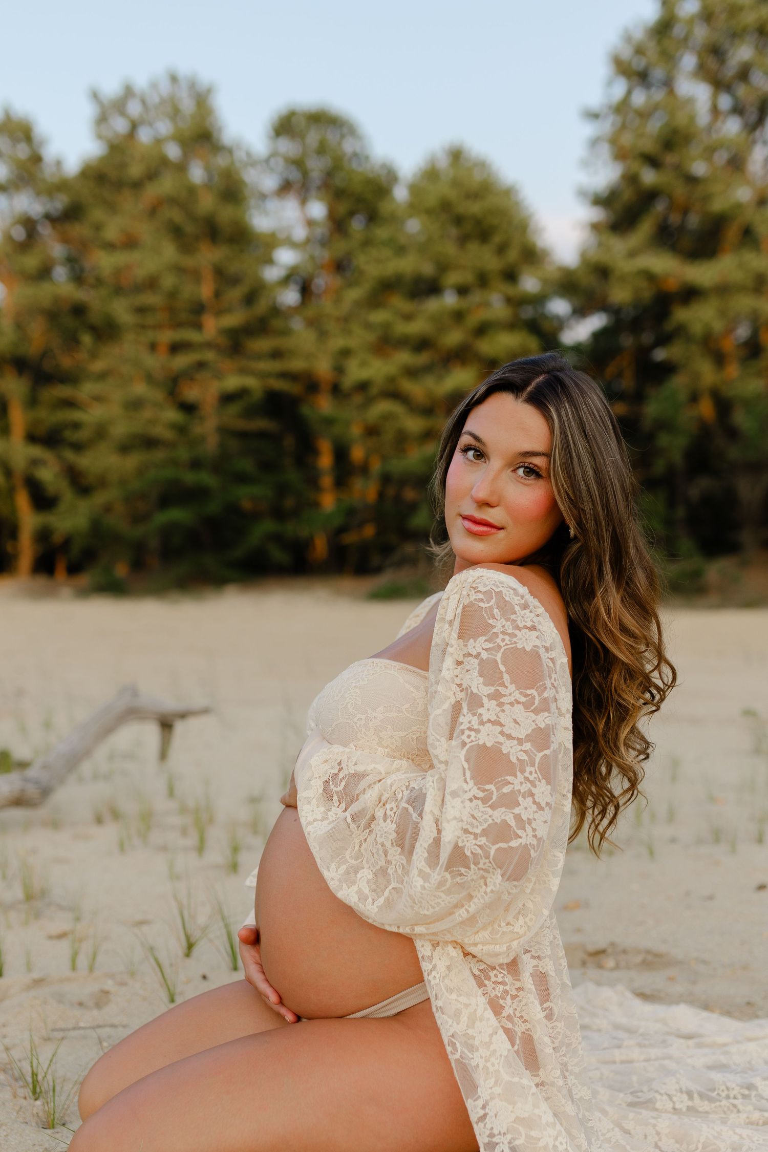 Pregnant woman in lace maternity robe sits on sand dunes surrounded by pine trees at sunset.