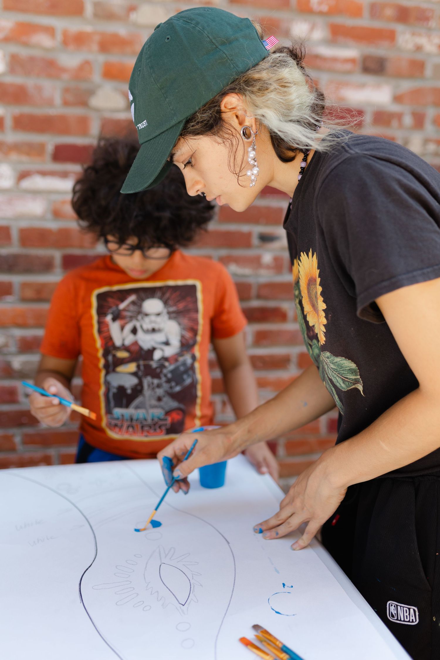 Woman in green cap and child painting on large white paper against brick wall.
