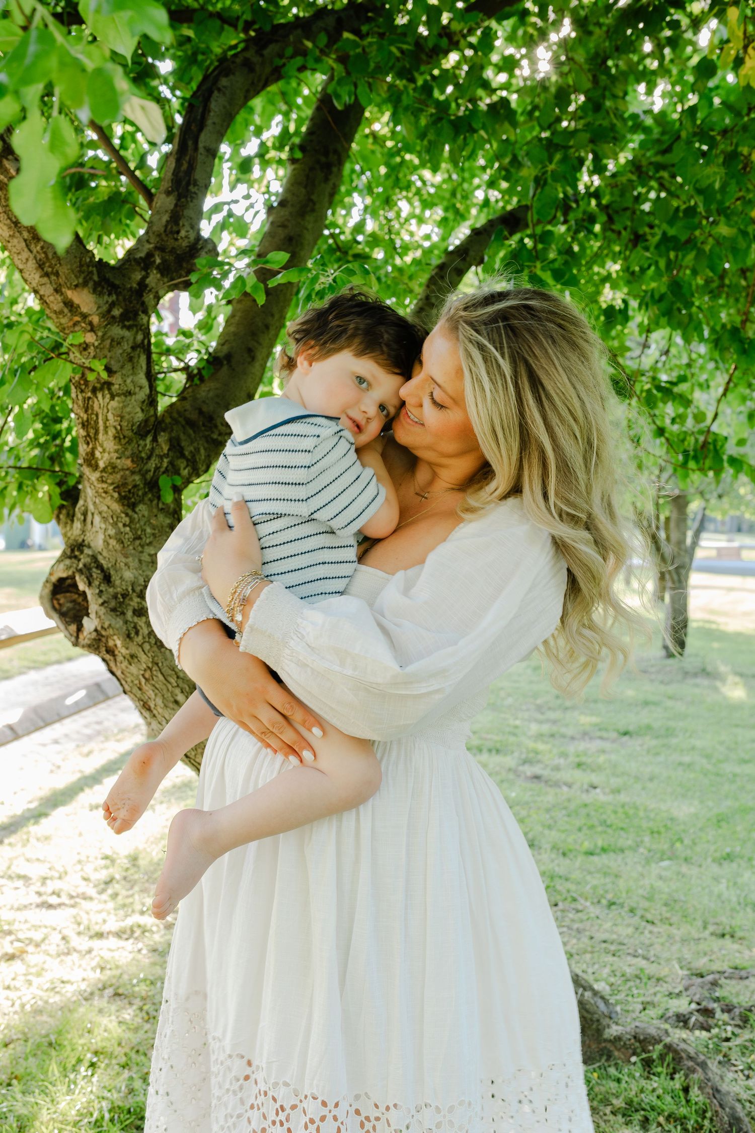 Blonde mother in white lace dress kissing her toddler son under a large green tree outdoors.