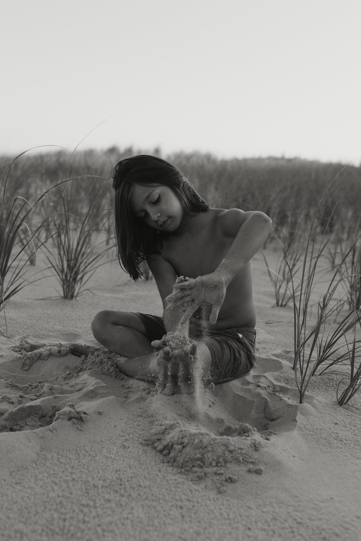 Black and white photo of a child sitting cross-legged on sand dunes letting sand fall through hands.