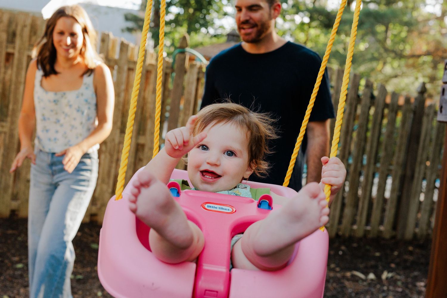 Happy toddler smiling in a pink Little Tikes swing with parents in a backyard.