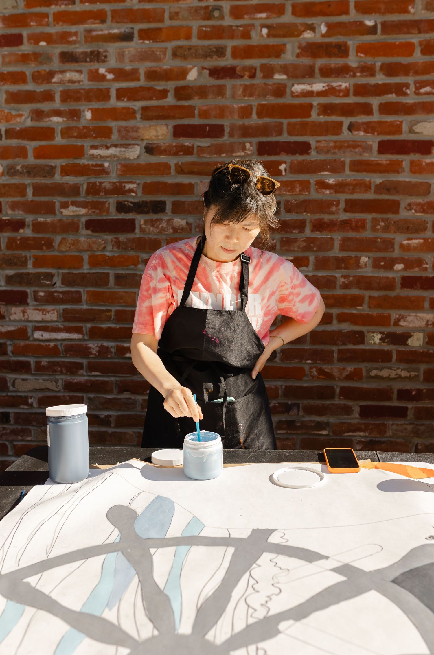 Artist in black apron painting blue design on large canvas outdoors against red brick wall.
